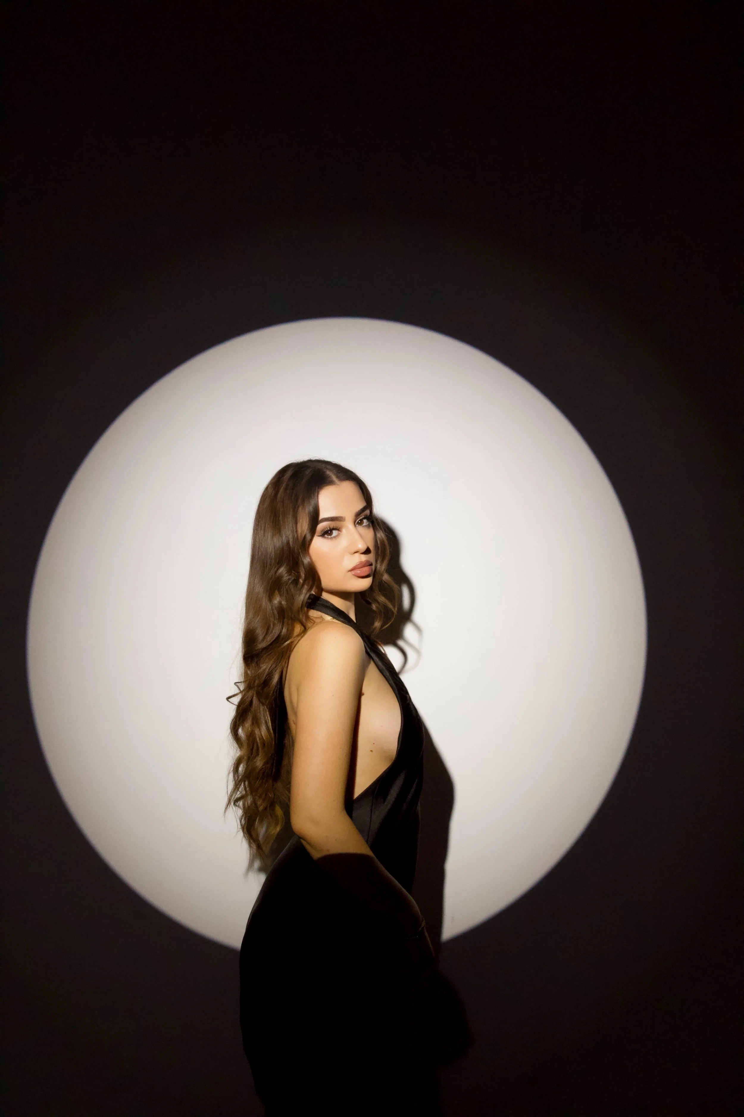 A woman with long, wavy brown hair and makeup posing in front of a black background with a white circular spotlight.