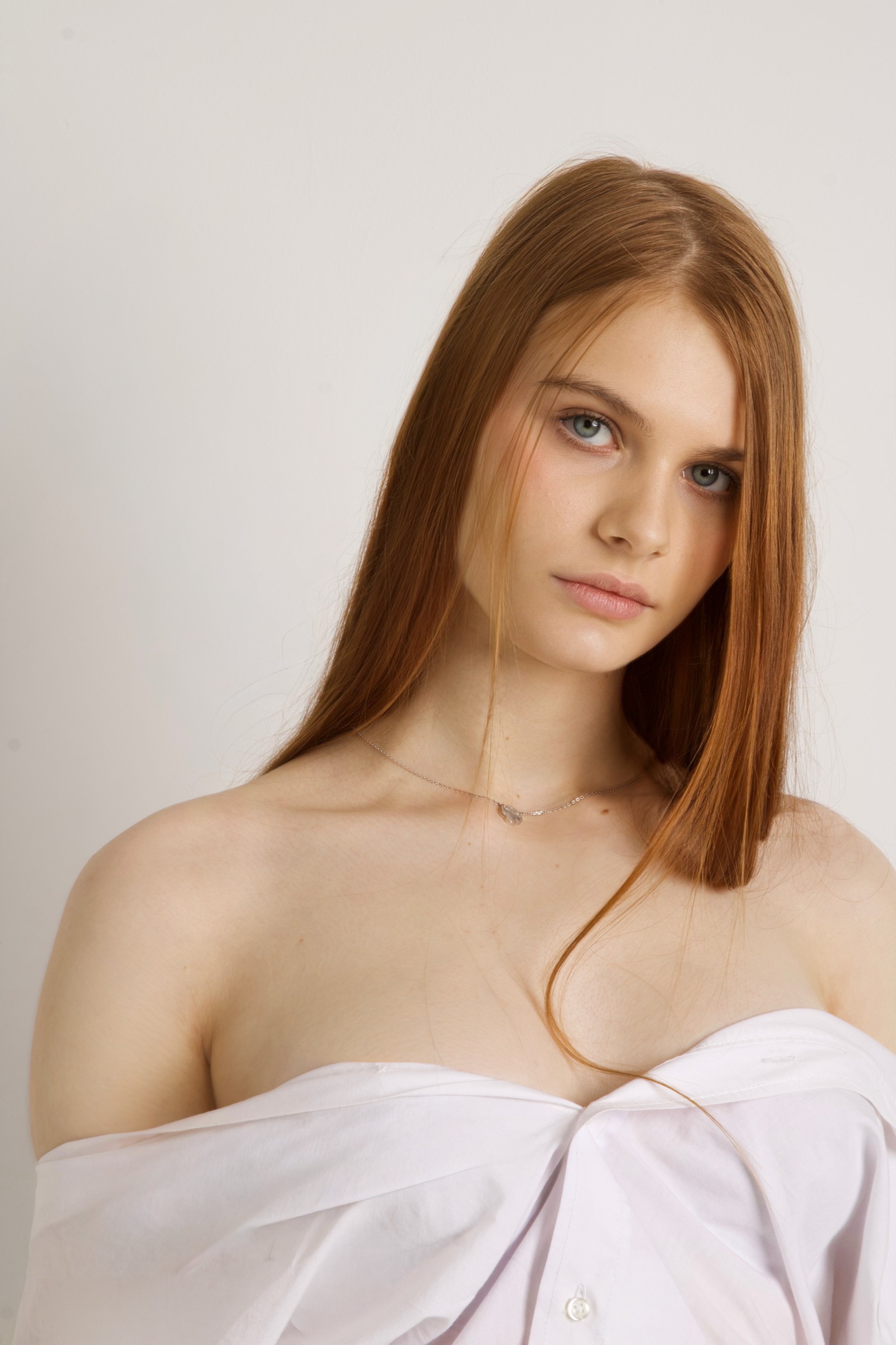 A young woman with long red hair, blue eyes, and fair skin, wearing a white off-shoulder top and a delicate necklace, standing against a plain white background.