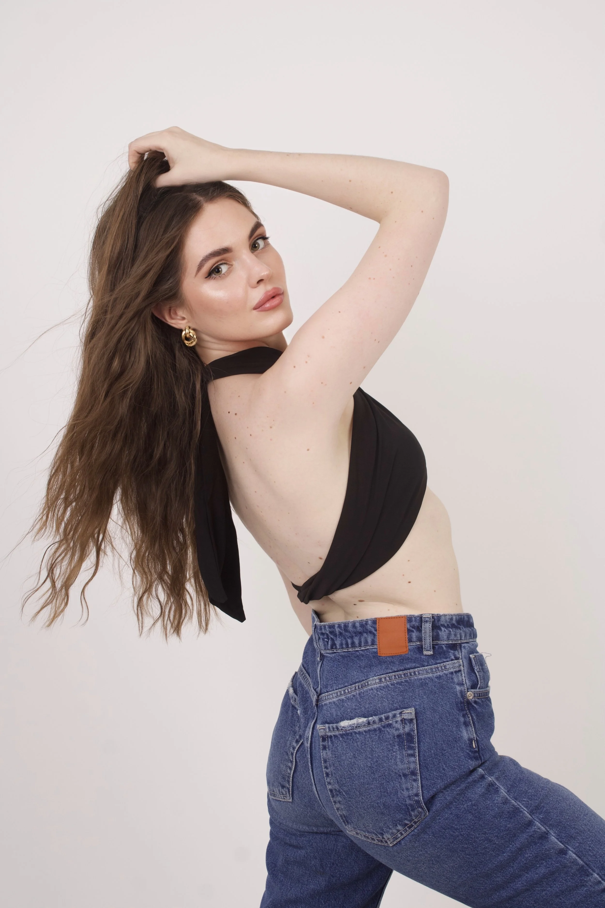 A young woman with long wavy hair posing against a plain white background. She is wearing hoop earrings, a black sleeveless top, and high-waisted blue jeans.