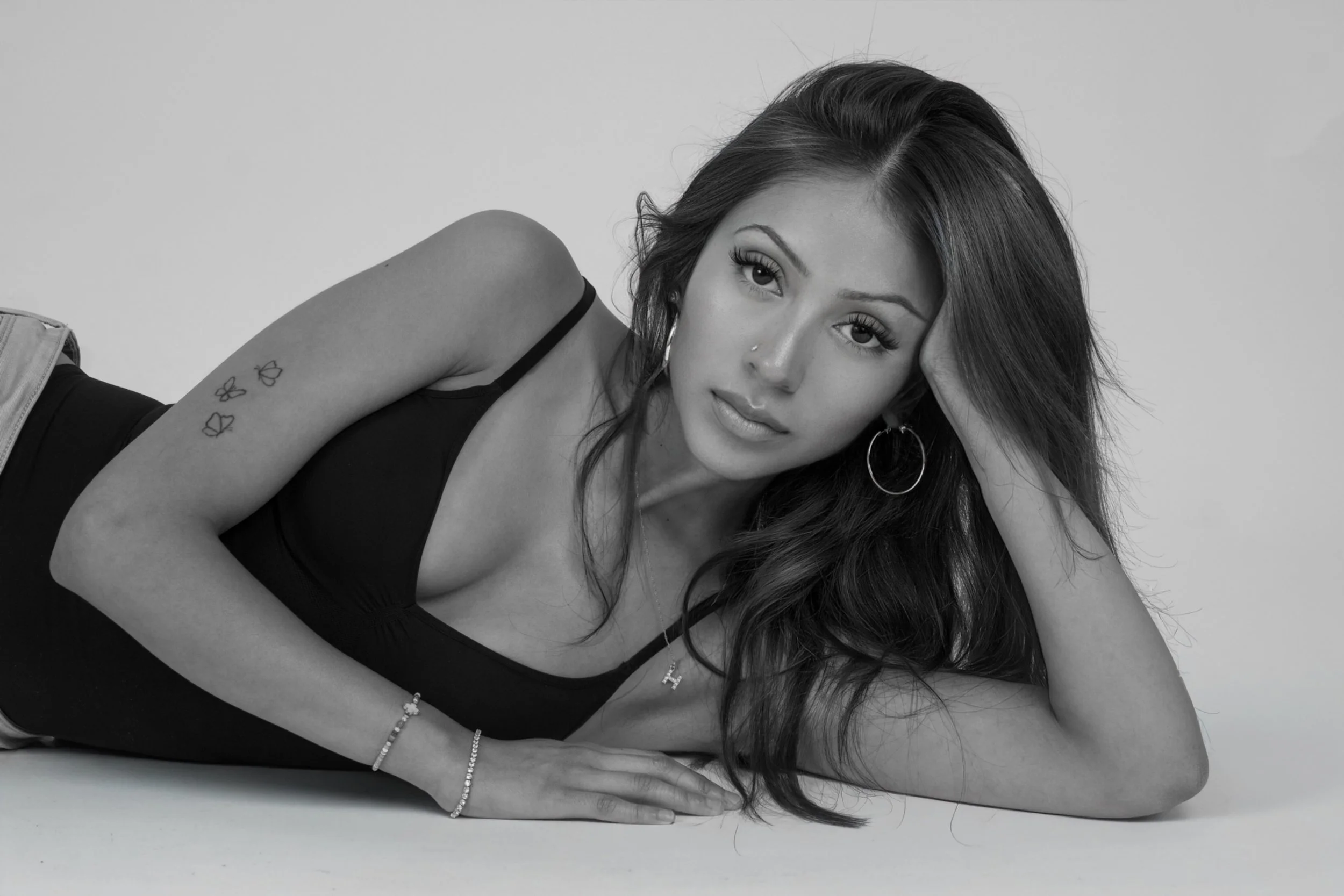 A black and white photo of a woman lying on her side with her head resting on her hand, wearing a black tank top and jewelry, looking at the camera.