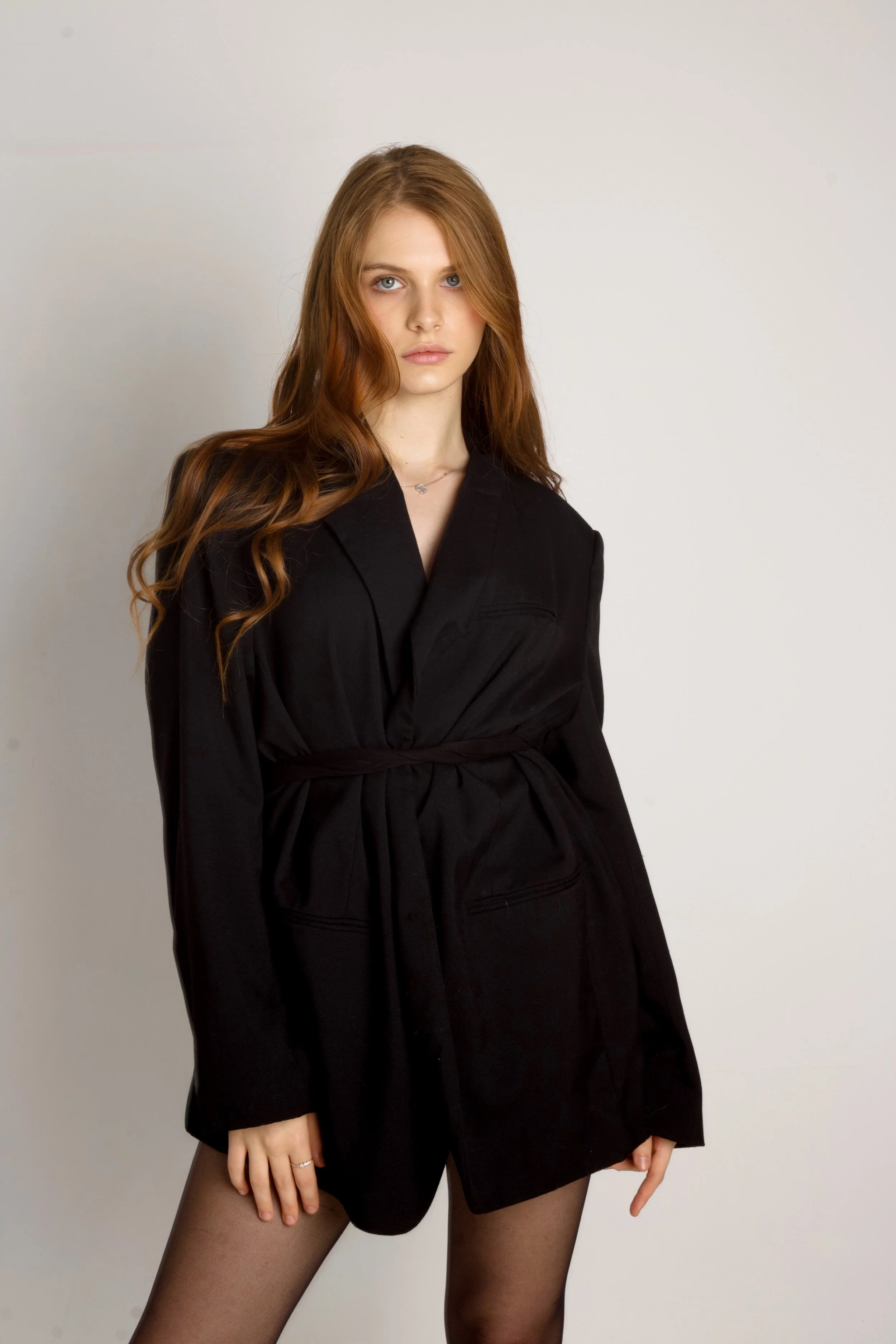 A young woman with long, wavy red hair and blue eyes, wearing a black jacket and black tights, standing against a plain light-colored background.