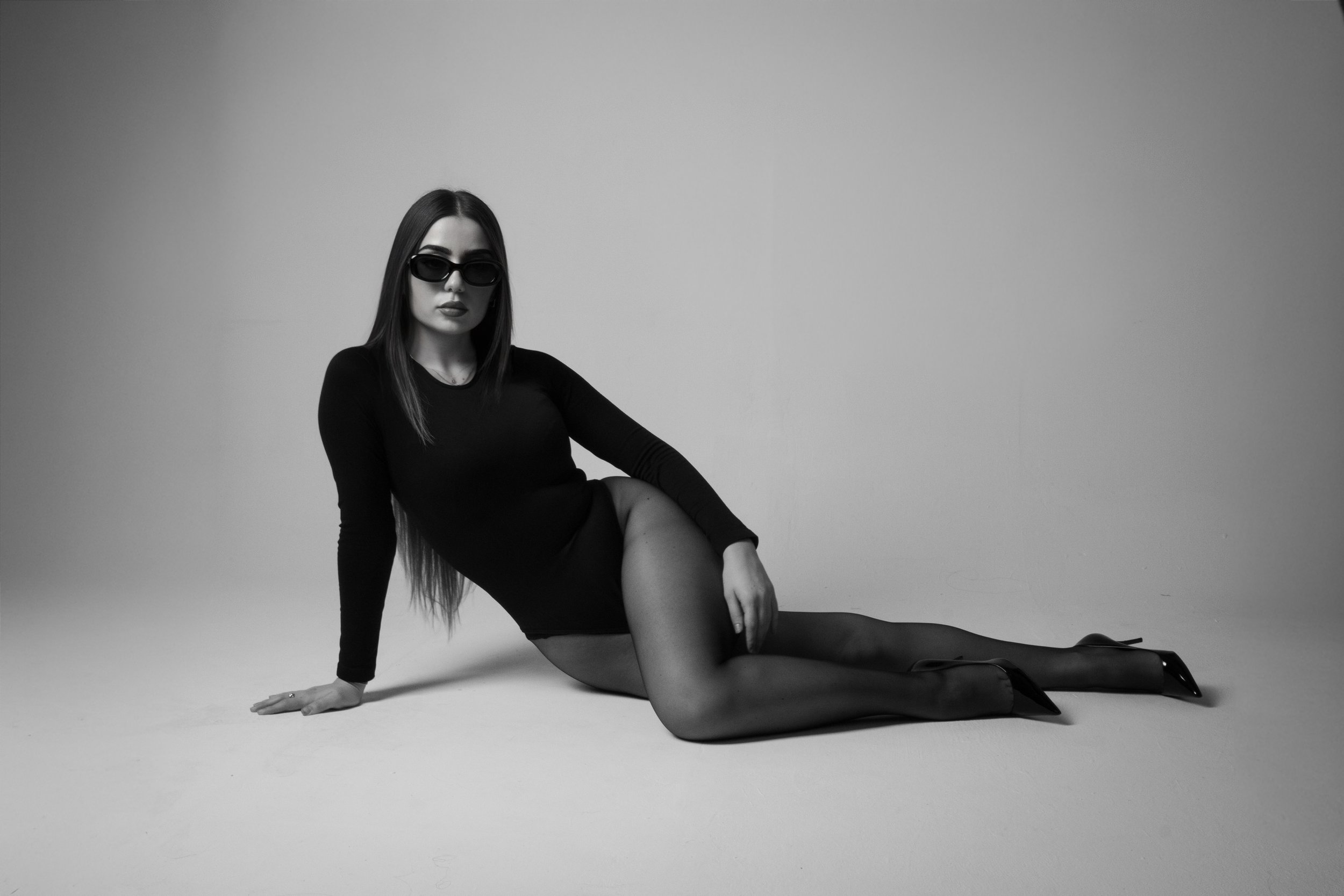 Black and white photo of a woman with long hair, wearing sunglasses, a black long-sleeve dress, sheer tights, and high heels, sitting on the floor with one leg extended and the other bent.