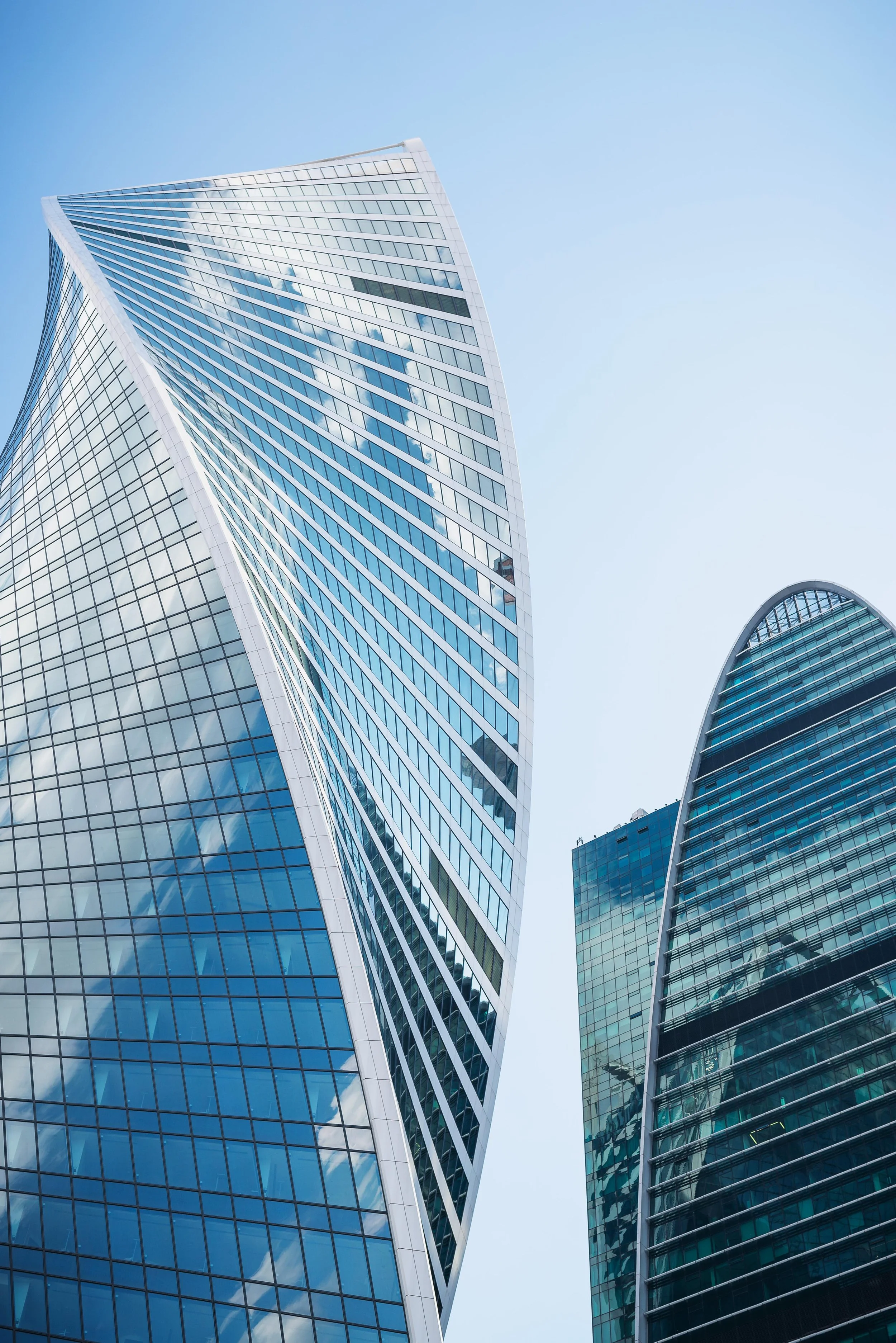 Two modern glass skyscrapers with reflections of clouds and sky, viewed from below against a pale blue sky.