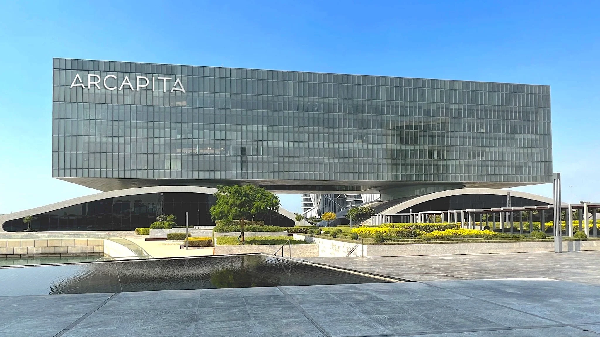 Modern building with the sign 'ARCAPITA' on top, glass facade, landscaped foreground with trees, plants, and water feature, under a clear blue sky.