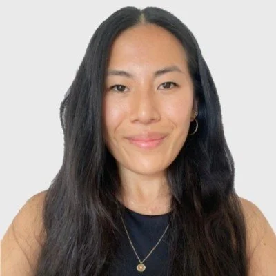 A woman with long black hair, wearing a black top, tan cardigan, and gold necklace, smiling at the camera against a light gray background.