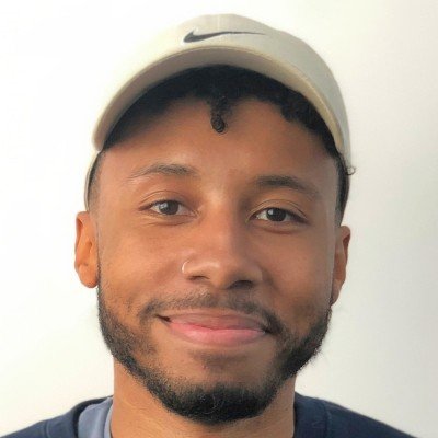 A young man with a beard and mustache wearing a beige Nike cap and a dark shirt, smiling against a plain light-colored wall.