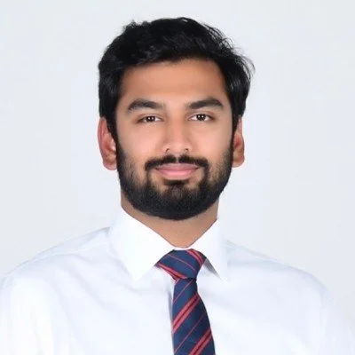 Portrait of a young man with dark hair and a beard, wearing a white shirt and a striped tie, against a plain white background.