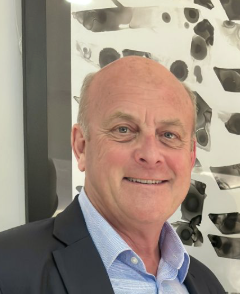 A smiling older man with a bald head and light skin, wearing a suit jacket and light blue dress shirt, standing indoors in front of a decorated wall.
