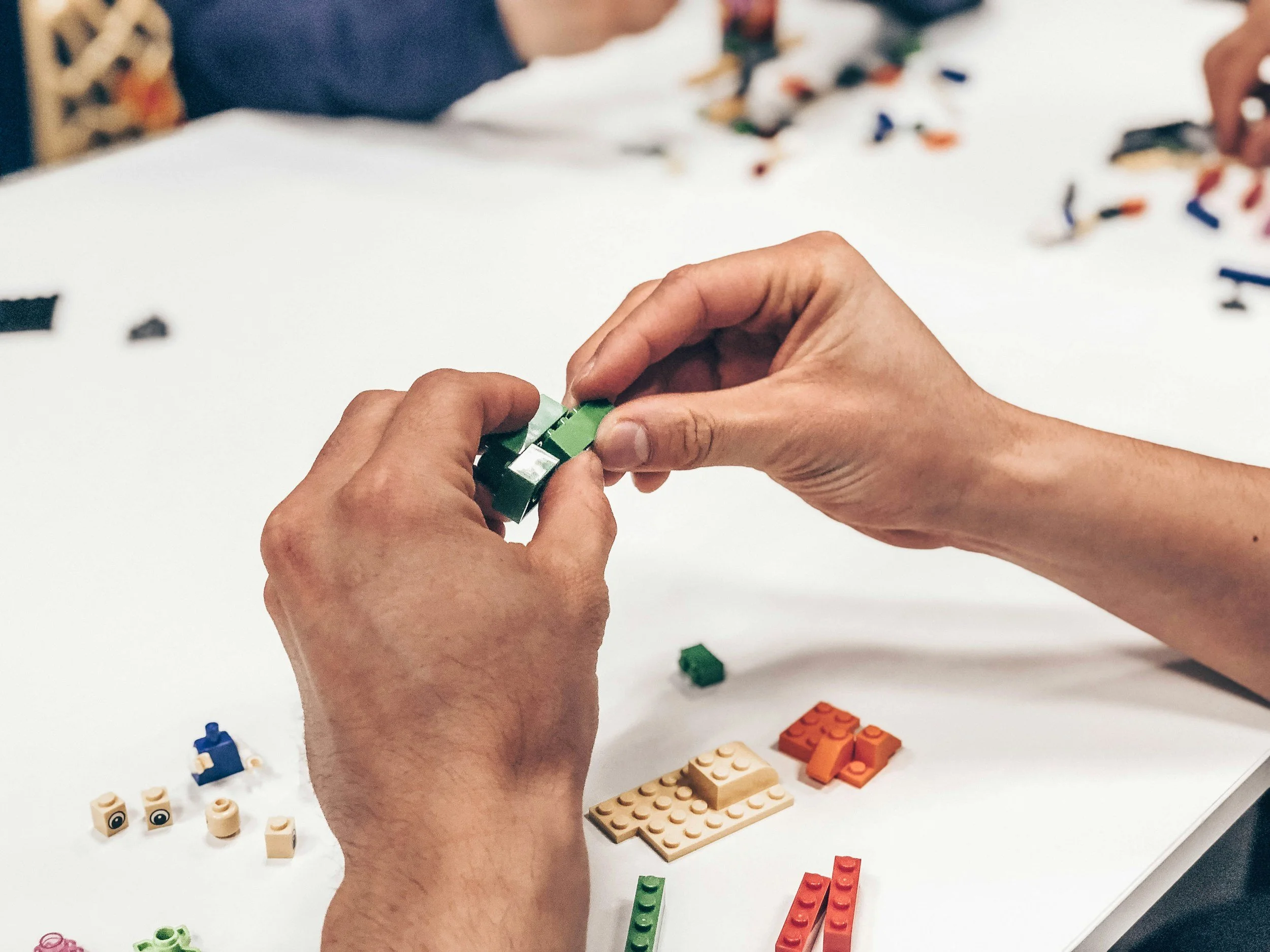 Hands assembling LEGO pieces on a white table, with various LEGO bricks scattered around.