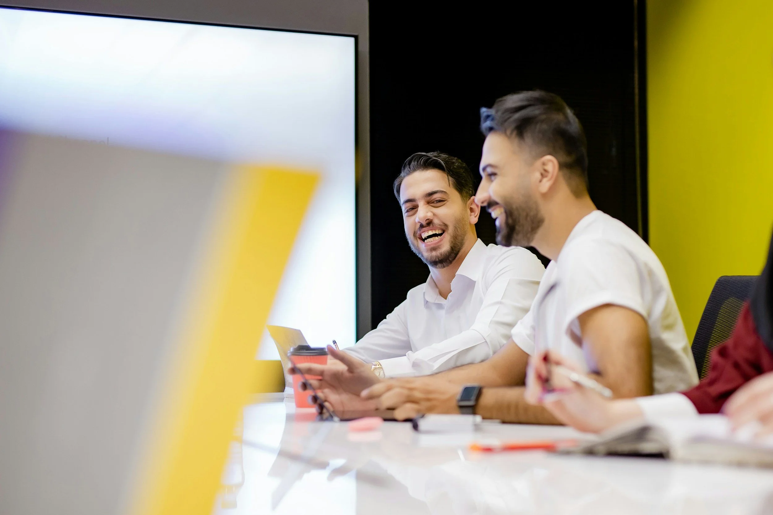Two young men in white shirts sitting at a conference table, laughing and talking, with laptops and notebooks in front of them, in a modern meeting room.
