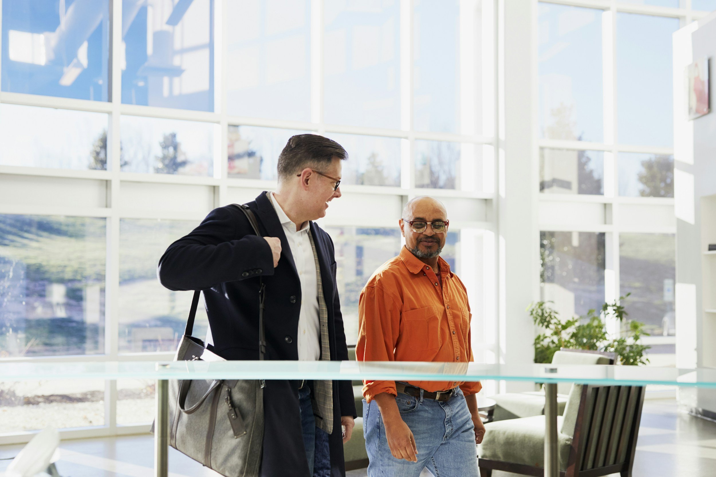 Two men walking and talking in a modern, brightly lit office with large windows and outdoor trees visible.