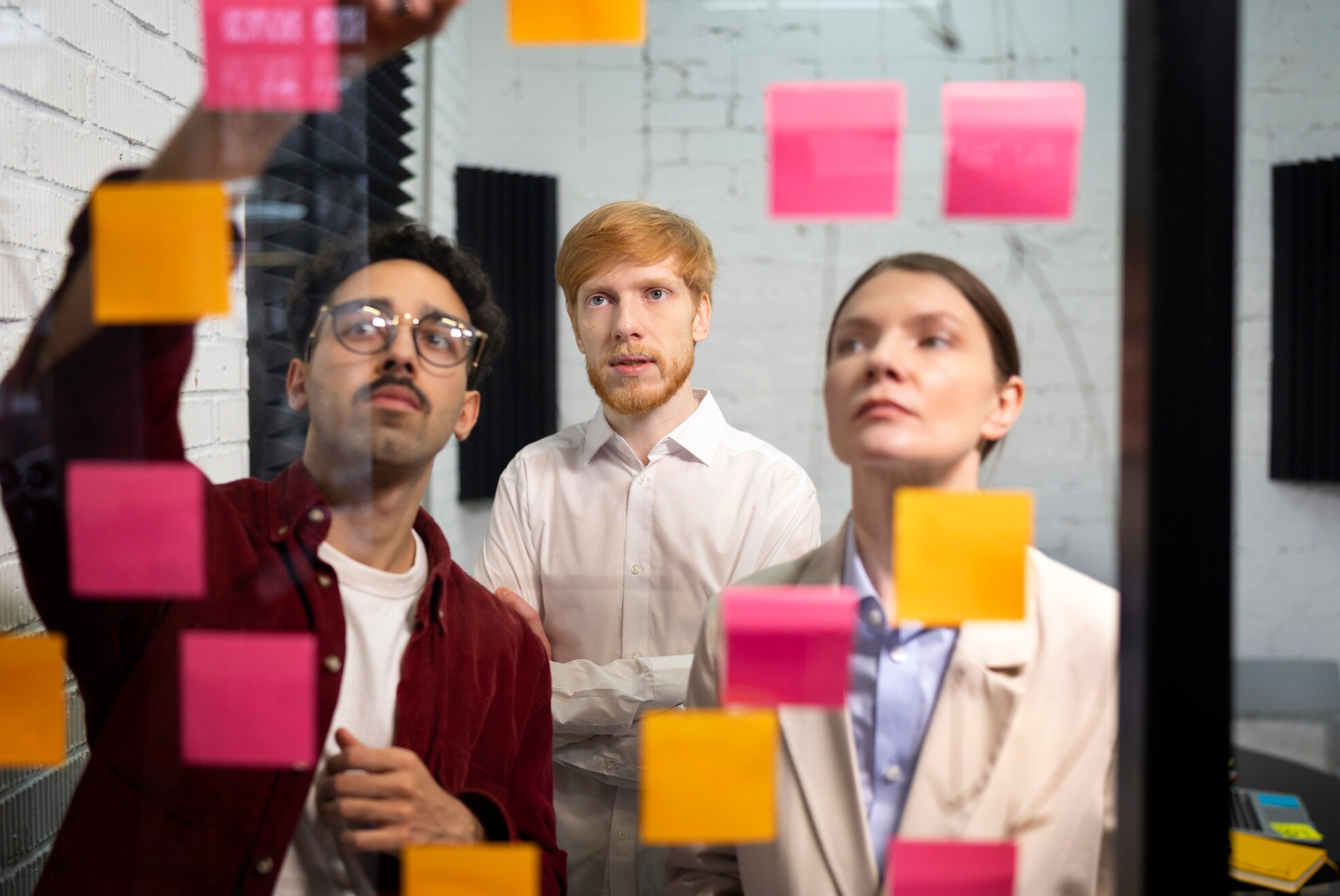 Three people examining pink and yellow sticky notes on a glass wall in a modern office or meeting room.