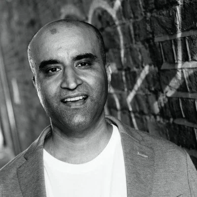 A man with a shaved head smiling, wearing a light-colored blazer over a white shirt, standing outdoors next to a brick wall with graffiti.