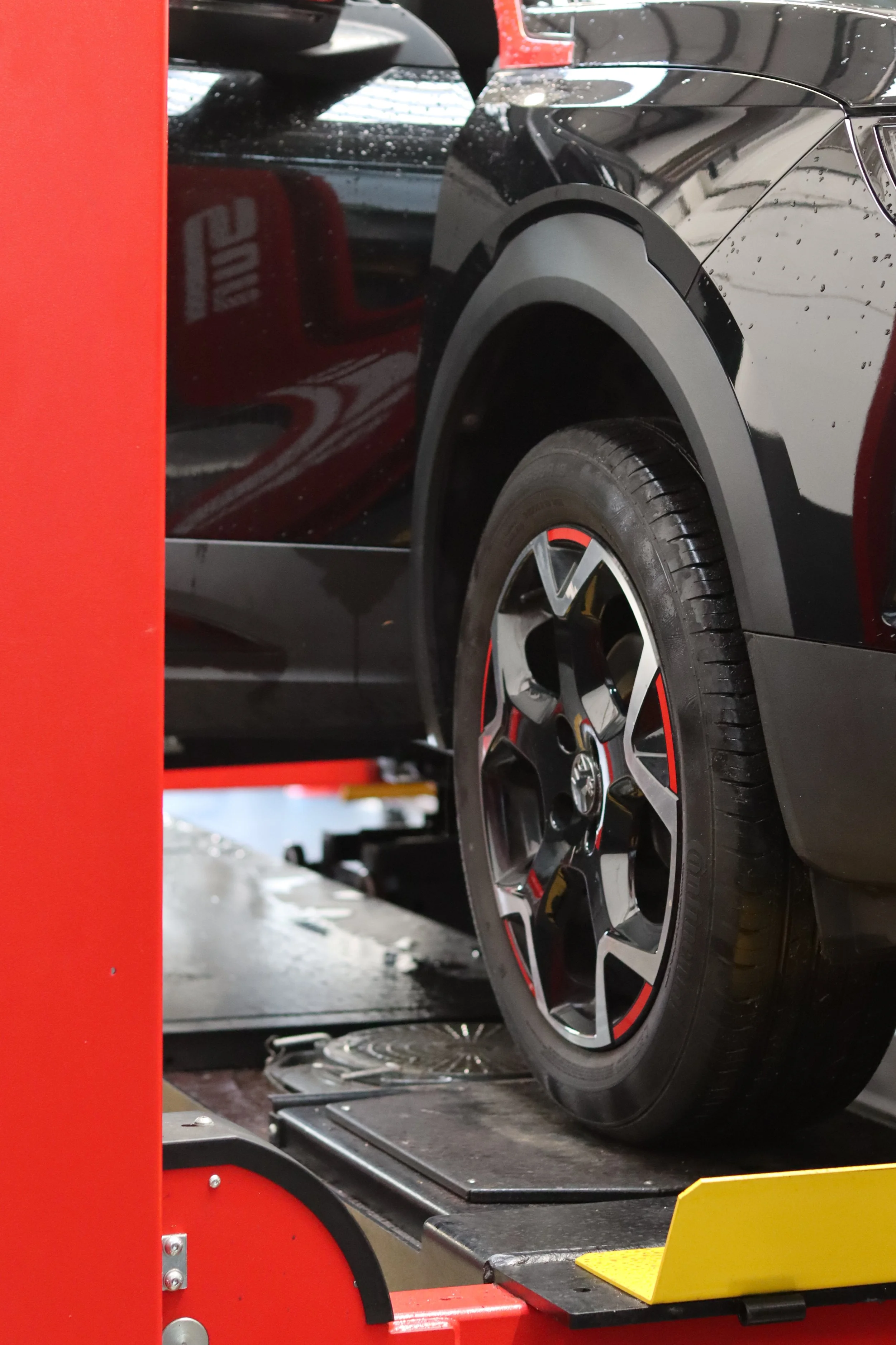 A black car with red and gray accents on the wheel rim is on a tilt machine at an auto repair or tire shop.