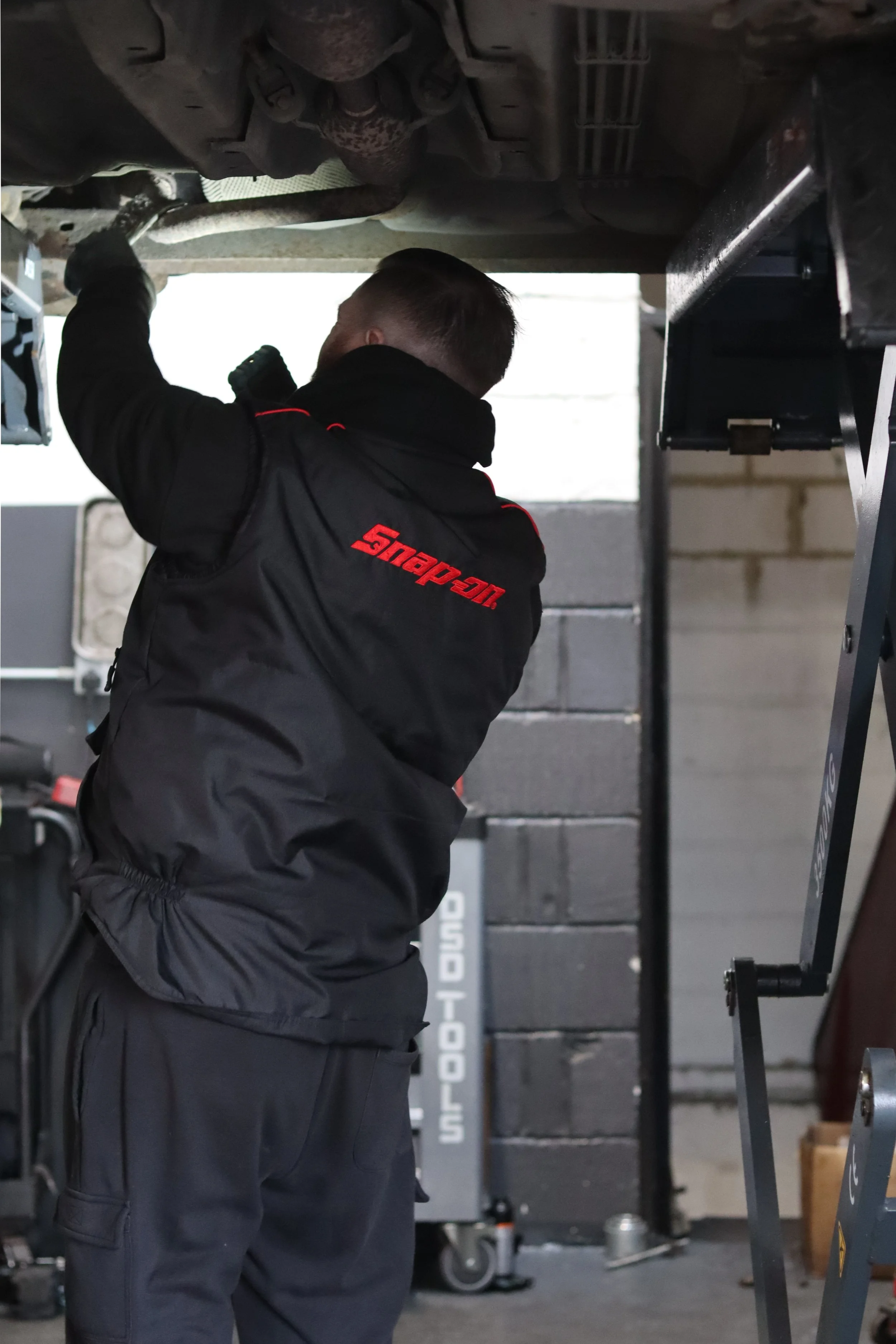 A mechanic working underneath a vehicle, wearing a black Snap-on jacket in a garage or workshop.