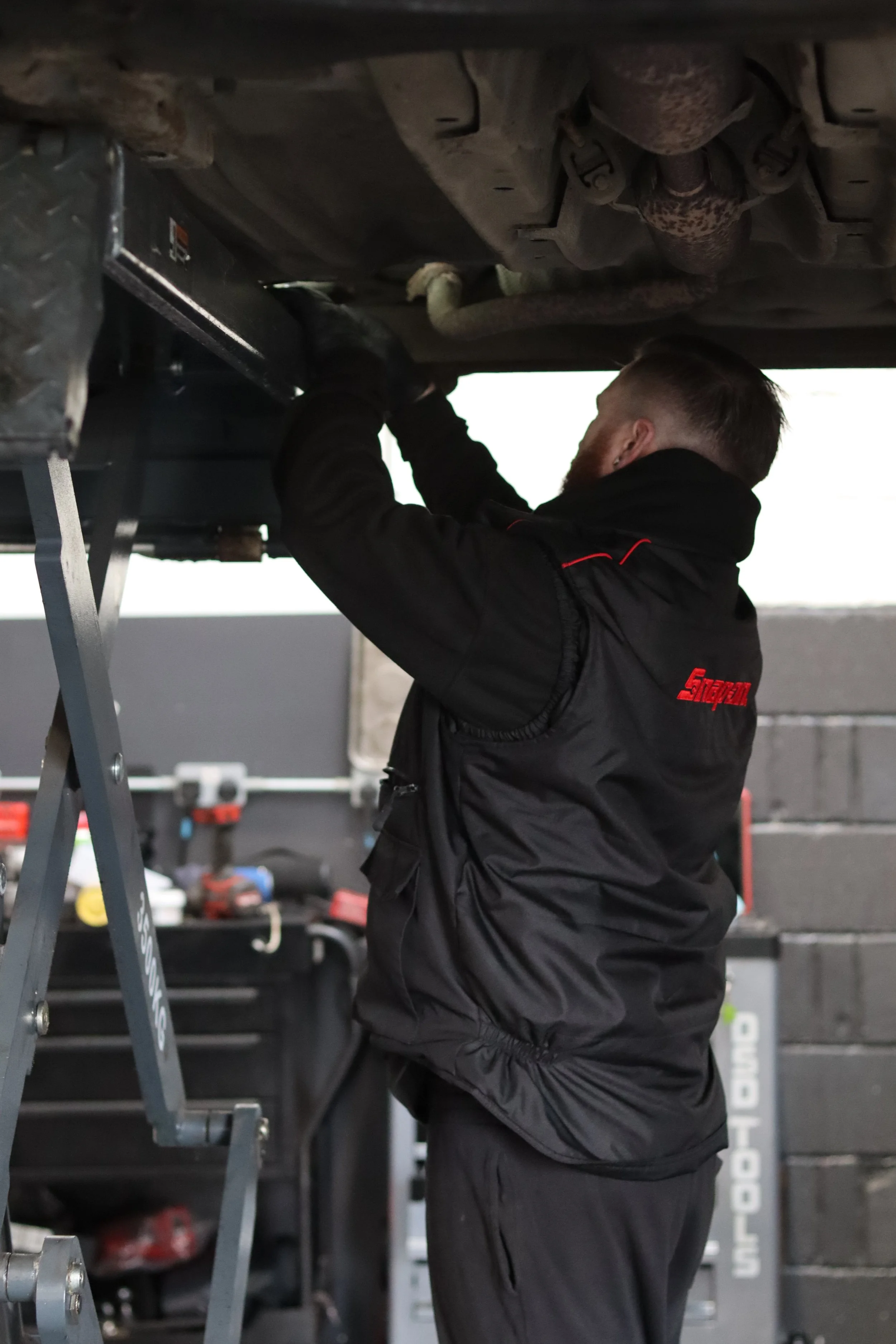 A man wearing a black jacket with red accents and gloves is working underneath a vehicle, using a lift or support. The vehicle's underside parts, including exhaust components, are visible.