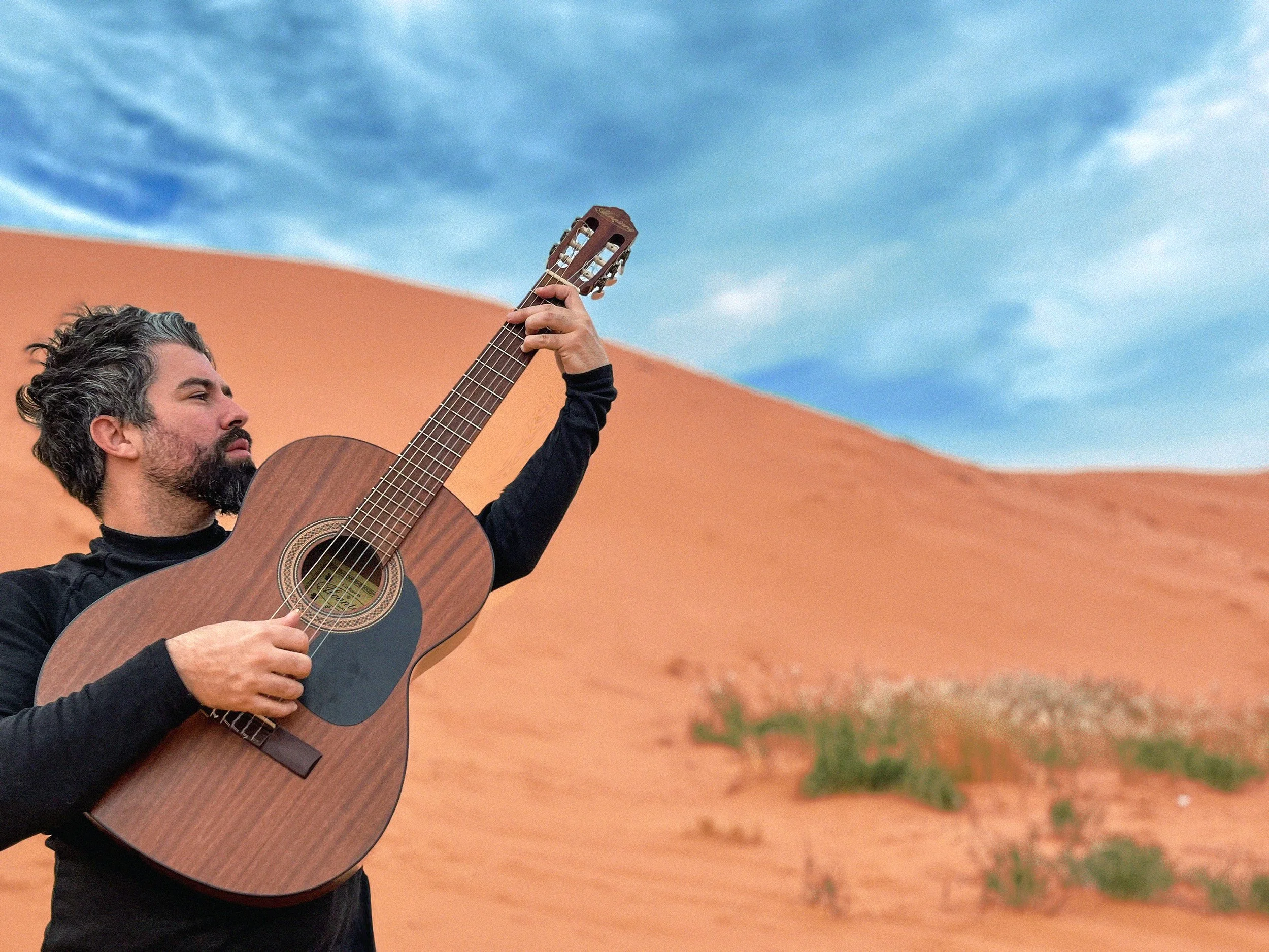 Un hombre tocando guitarra en un desierto con dunas y cielo azul con nubes.