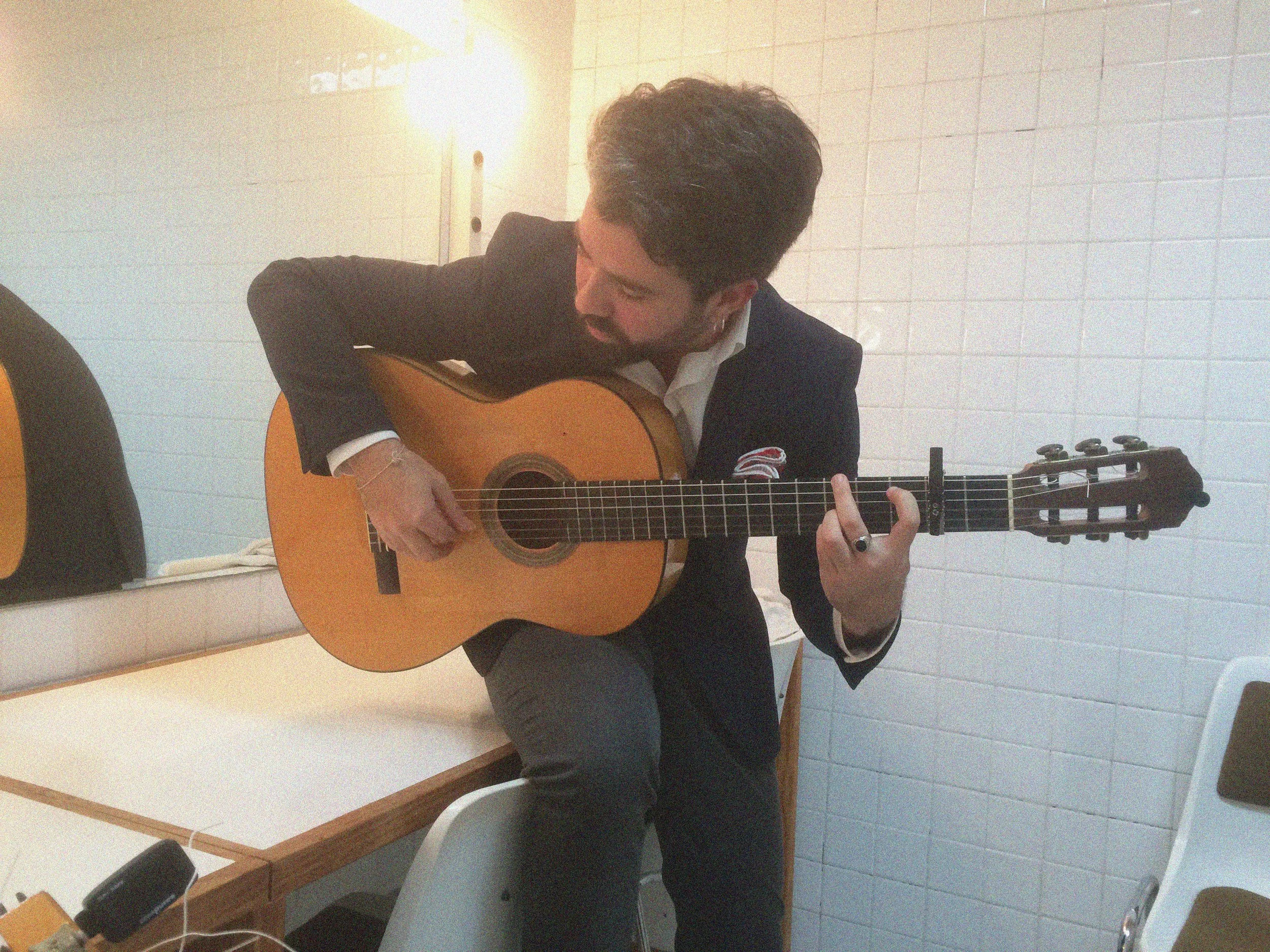Hombre tocando guitarra acústica en una sala con paredes blancas y luz cálida.