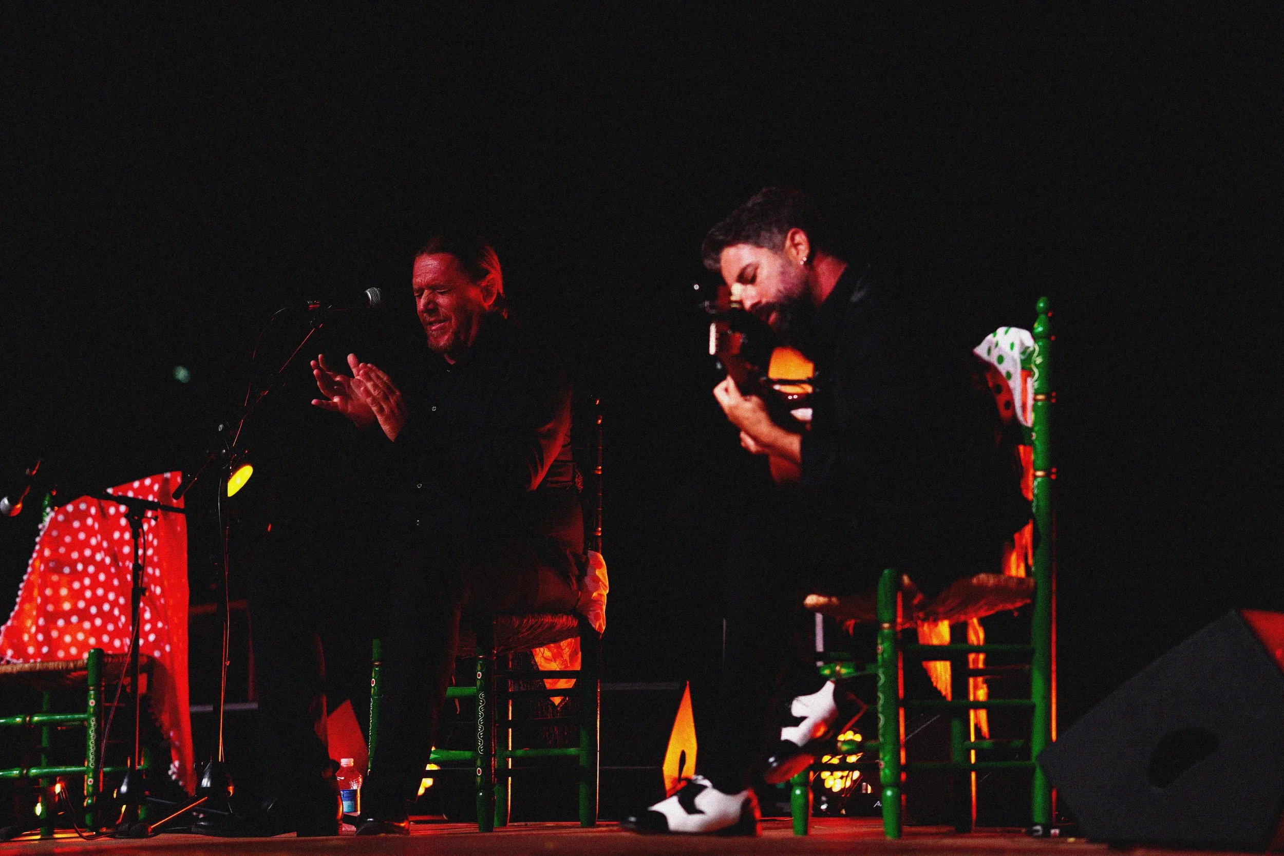 Dos hombres en un escenario en la noche, uno con los ojos cerrados y manos juntas, el otro tocando una guitarra, con sillas decoradas con tela de colores y luces focusing en ellos.