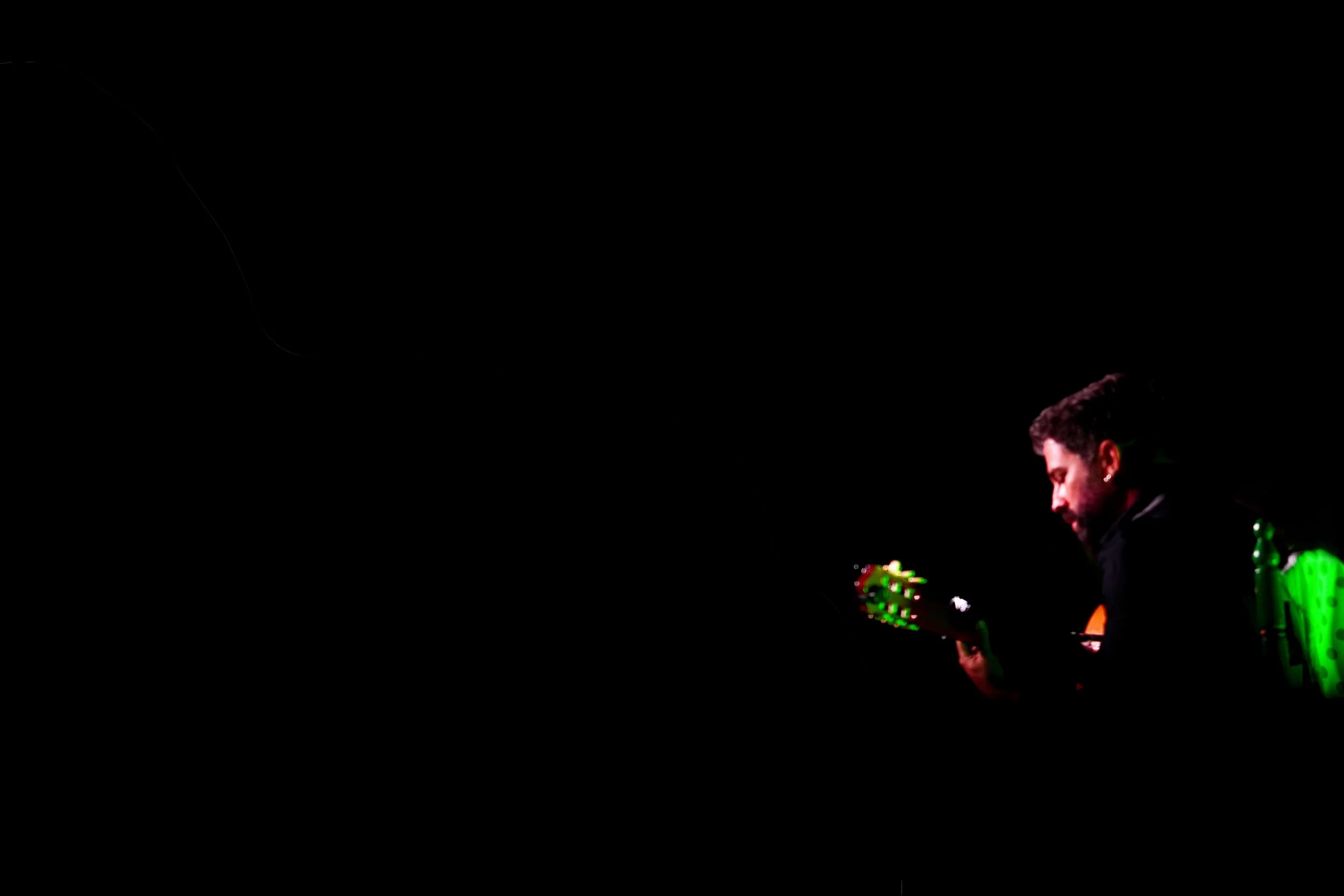 Hombre sentado en una silla, jugando un instrumento musical en un escenario oscuro con iluminación focalizada.