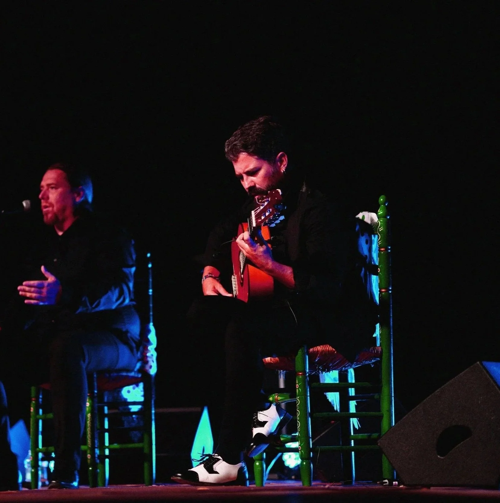 Dos hombres en un escenario, uno con guitarra y el otro posiblemente en una interpretación musical, en un entorno oscuro con iluminación teatral.