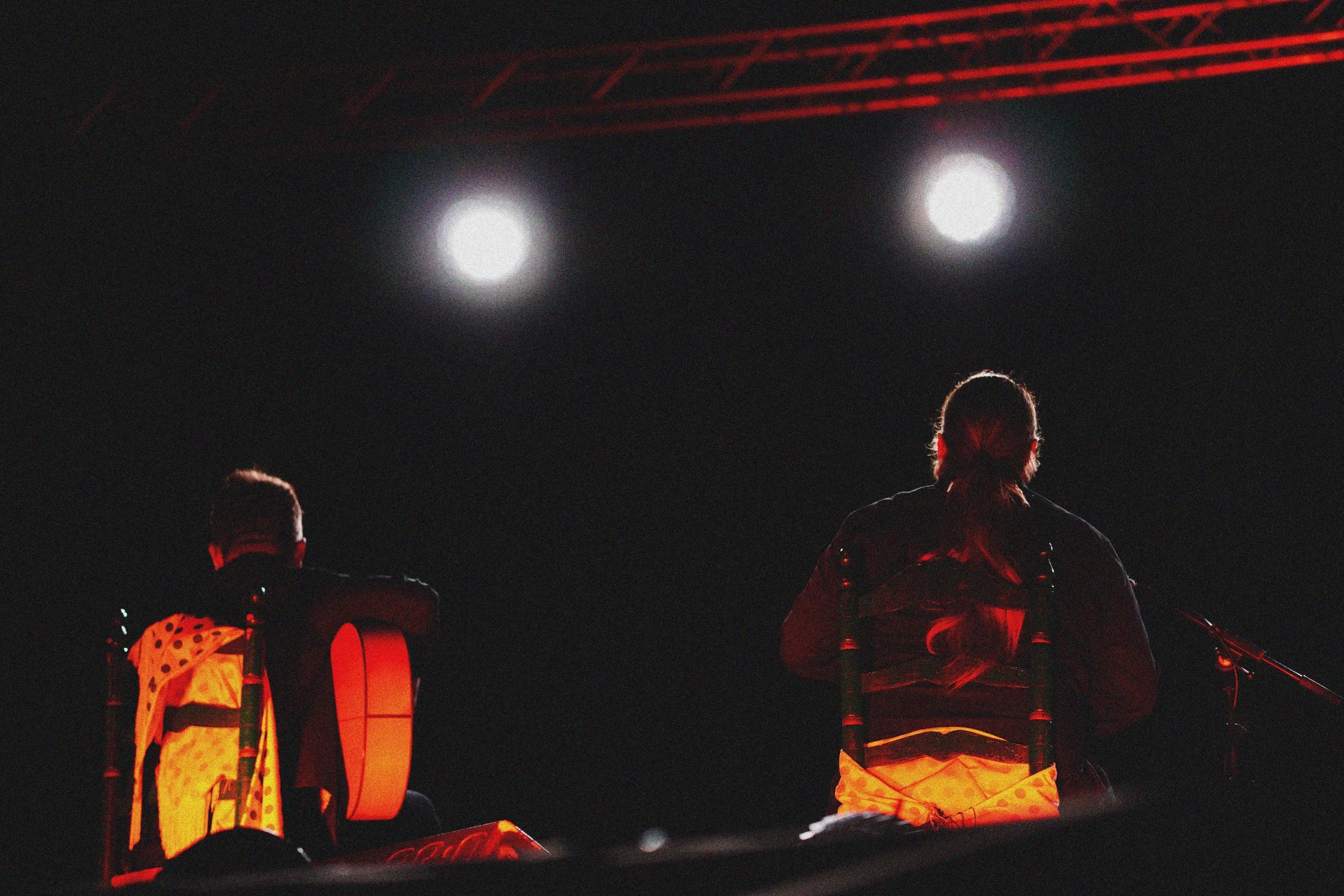 Dos personas sentadas frente a un micrófono en un escenario oscuro con luces blancas.