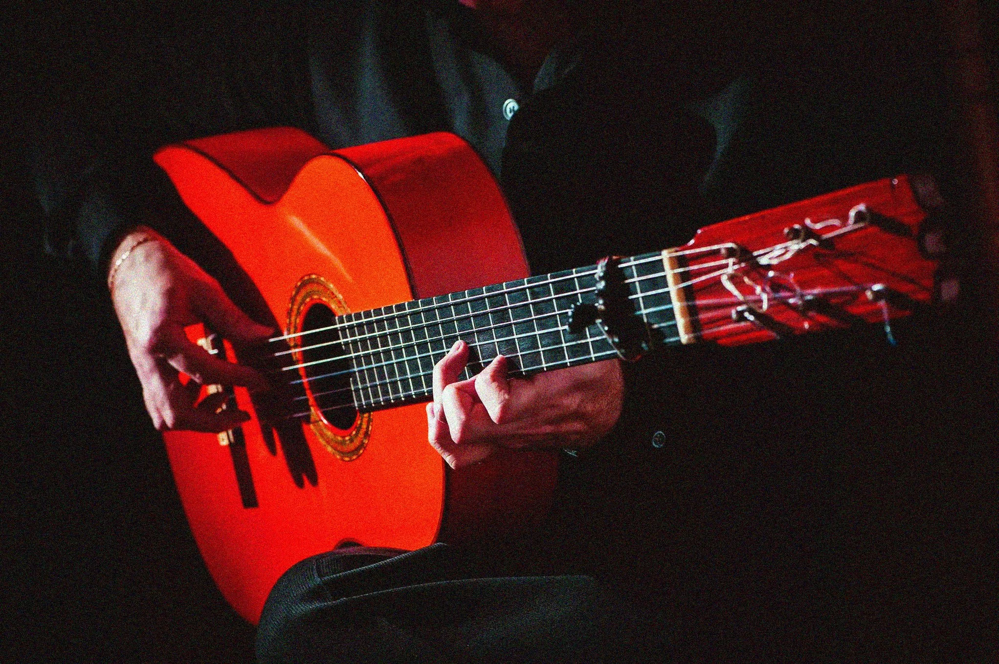 Persona tocando una guitarra acústica de color rojo en un ambiente oscuro.