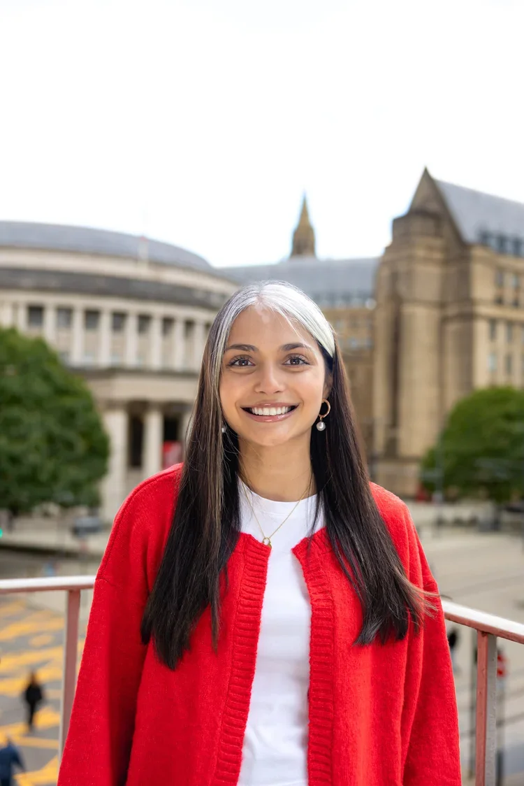 A smiling woman with long dark hair, wearing a red cardigan, white shirt, and earrings, standing outdoors with a historic building and trees in the background.