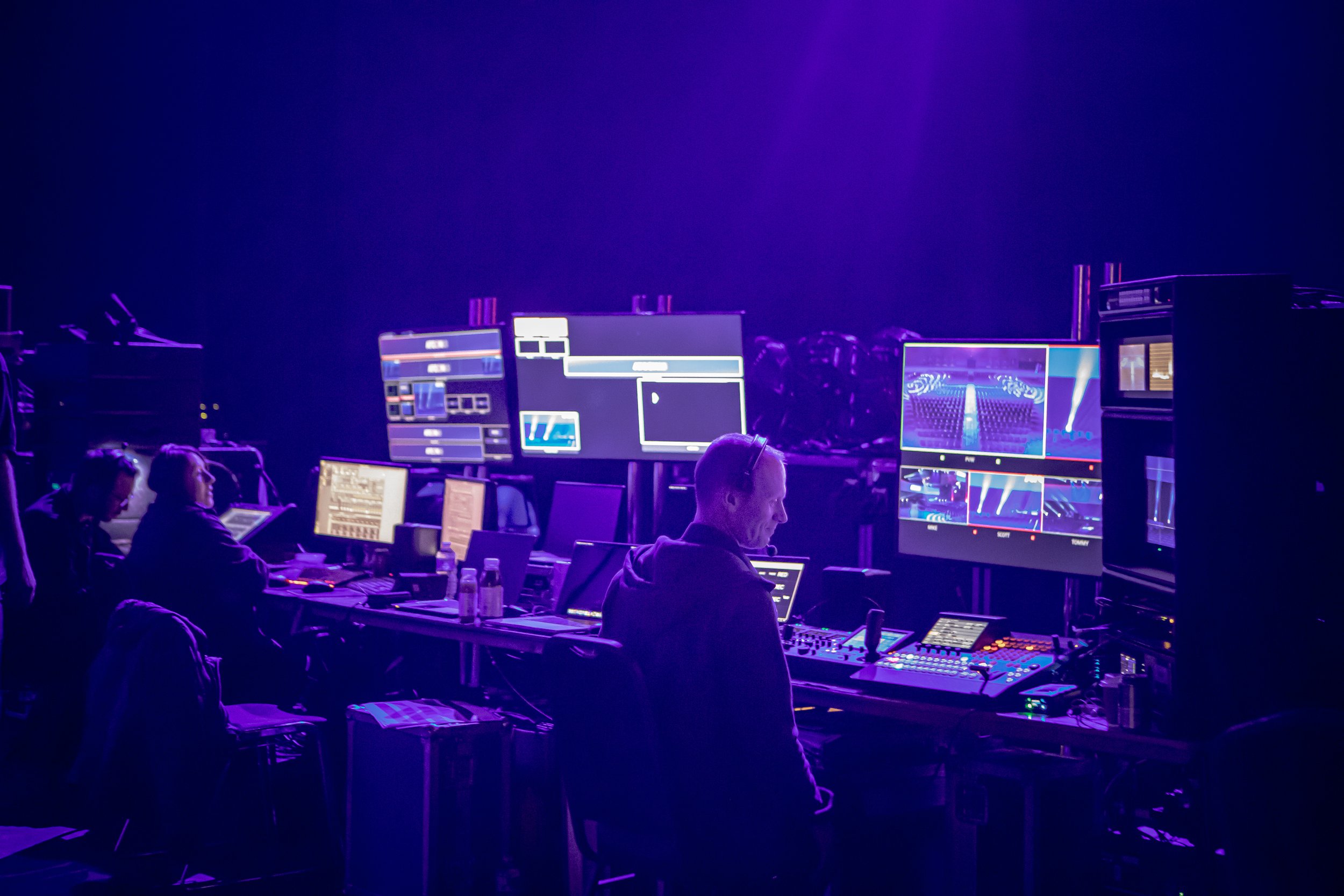 People working at a control room with multiple monitors showing stage lighting and video feeds, illuminated by purple stage lights