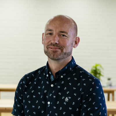 Lee Cuffe - A man with a beard and shaved head wearing a dark blue shirt with a small white pattern, standing indoors with a blurred background that includes potted plants.