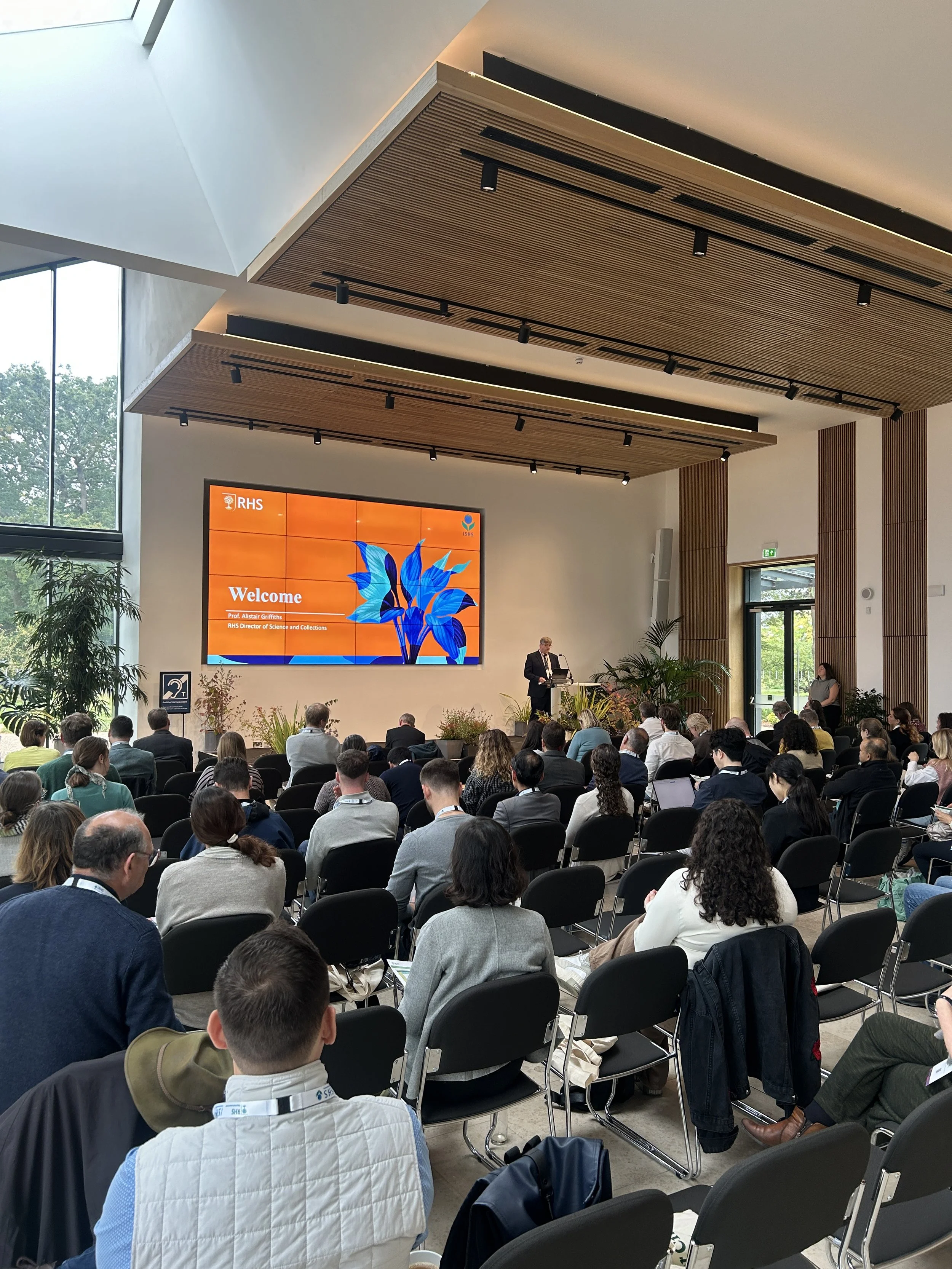 Conference room with a large audience attending a presentation, with a speaker at a podium and a large digital display screen showing the words 'Welcome' and 'RHS'.