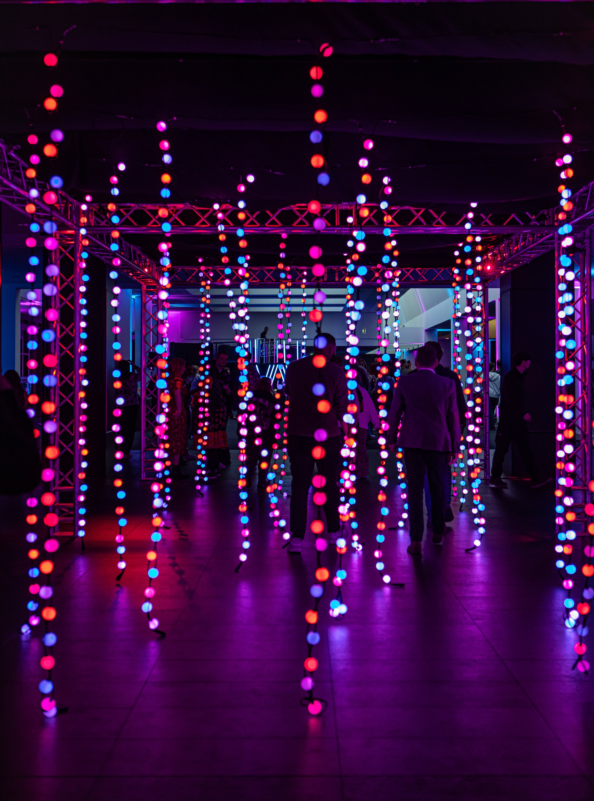 People walking through an art installation with hanging colorful LED lights in a darkened room.