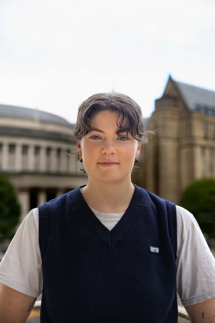 Young woman with short curly hair, wearing a white t-shirt and navy vest, standing outdoors in front of historic buildings and greenery.