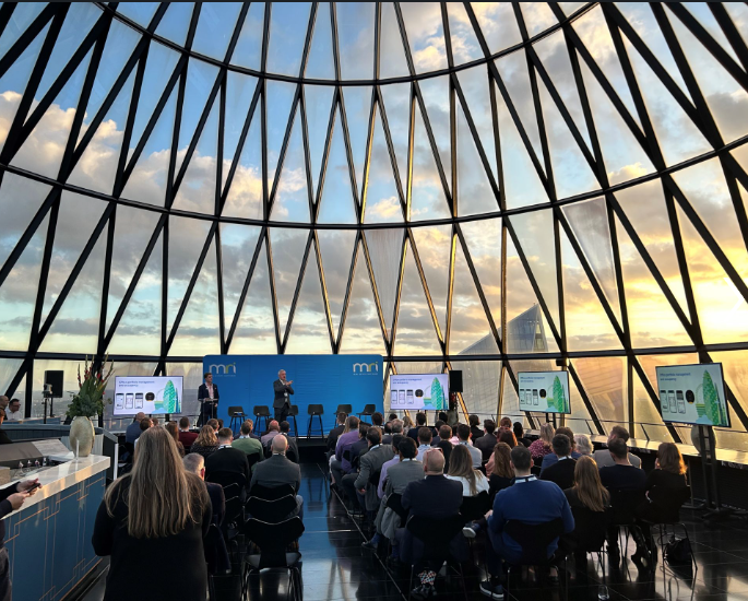 A presentation or conference taking place inside a glass dome with a city skyline view at sunset. Attendees sit facing a stage with speakers and large screens displaying slides.