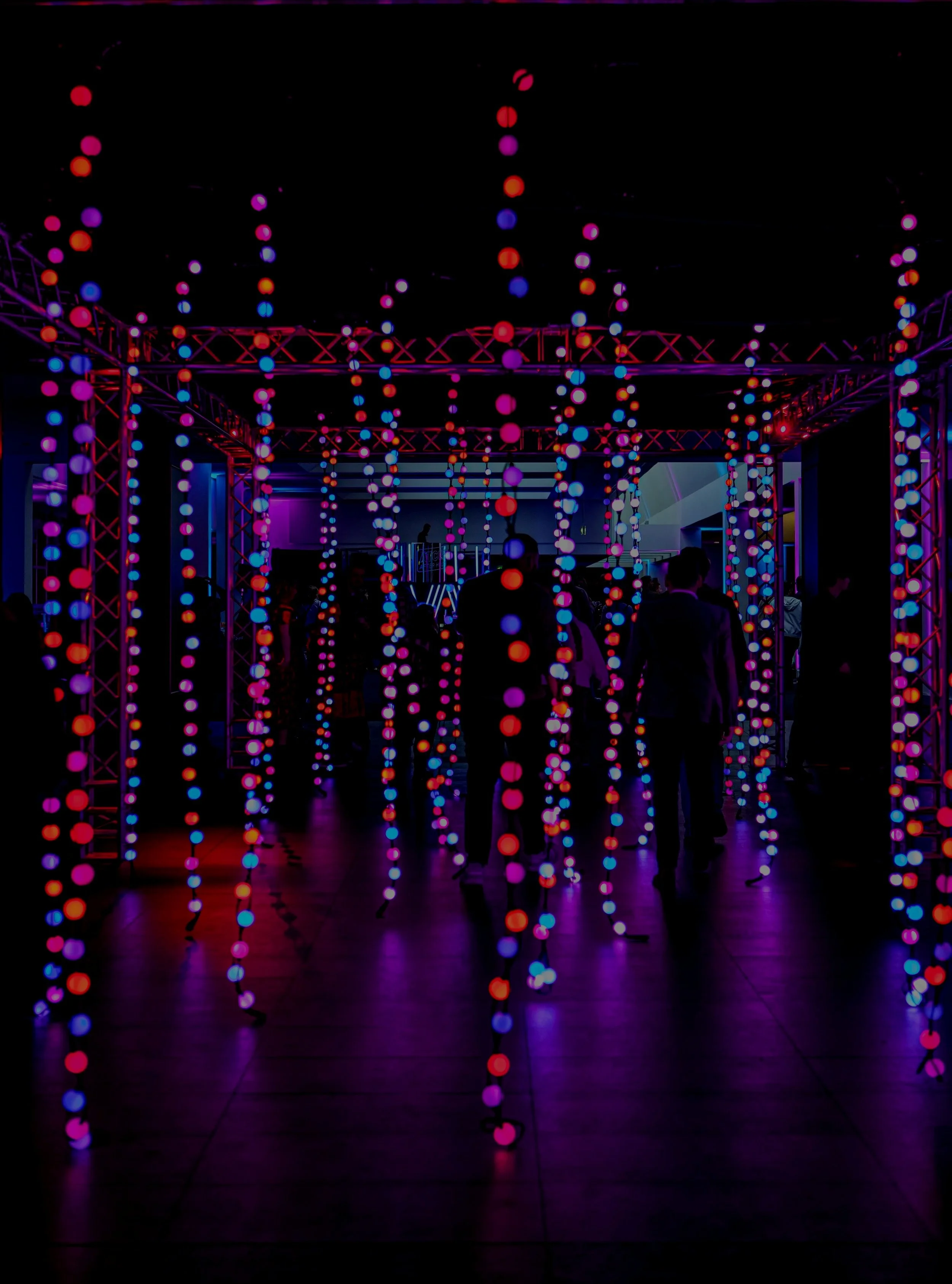 Dark room decorated with hanging colorful string lights in pink, purple, red, and blue, creating a vibrant atmosphere with silhouettes of people visible.