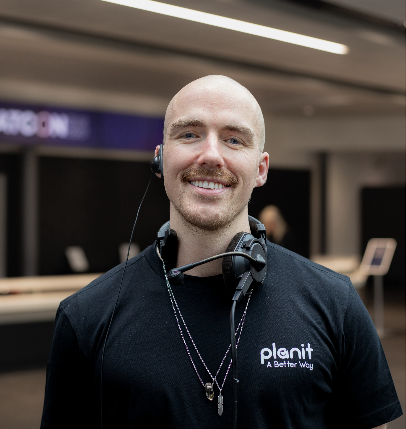 A smiling man with a shaved head and a mustache wearing a black t-shirt with the logo 'planit A Better Way.' He has a headset with a microphone around his neck and is wearing two necklaces. The background appears to be an indoor tech or office environment.