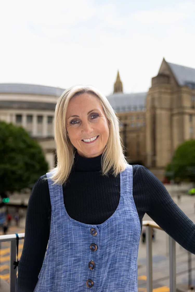 A smiling woman with blonde hair wearing a black turtleneck and a blue plaid sleeveless dress standing outdoors in front of a historic building with a clock tower and trees.