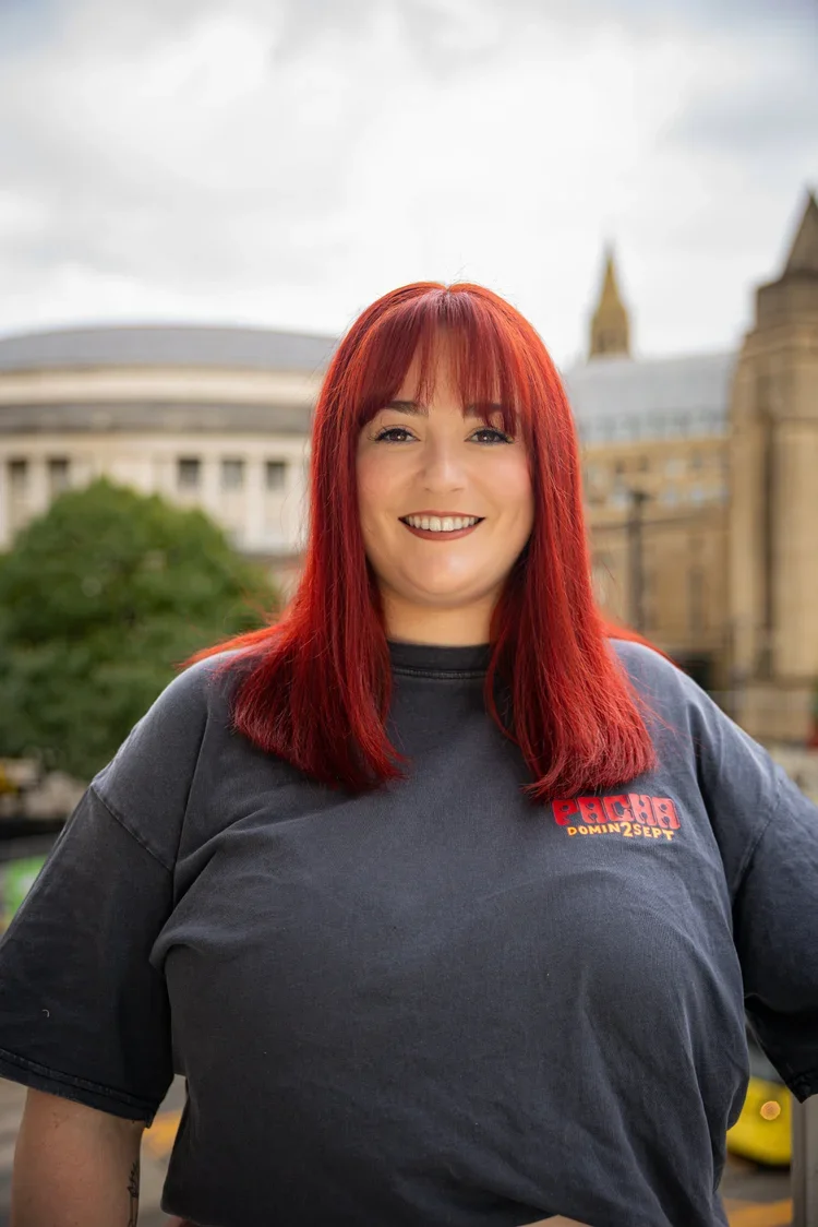 A smiling woman with red hair and a dark gray T-shirt standing outdoors in front of a cityscape with trees and historic buildings.