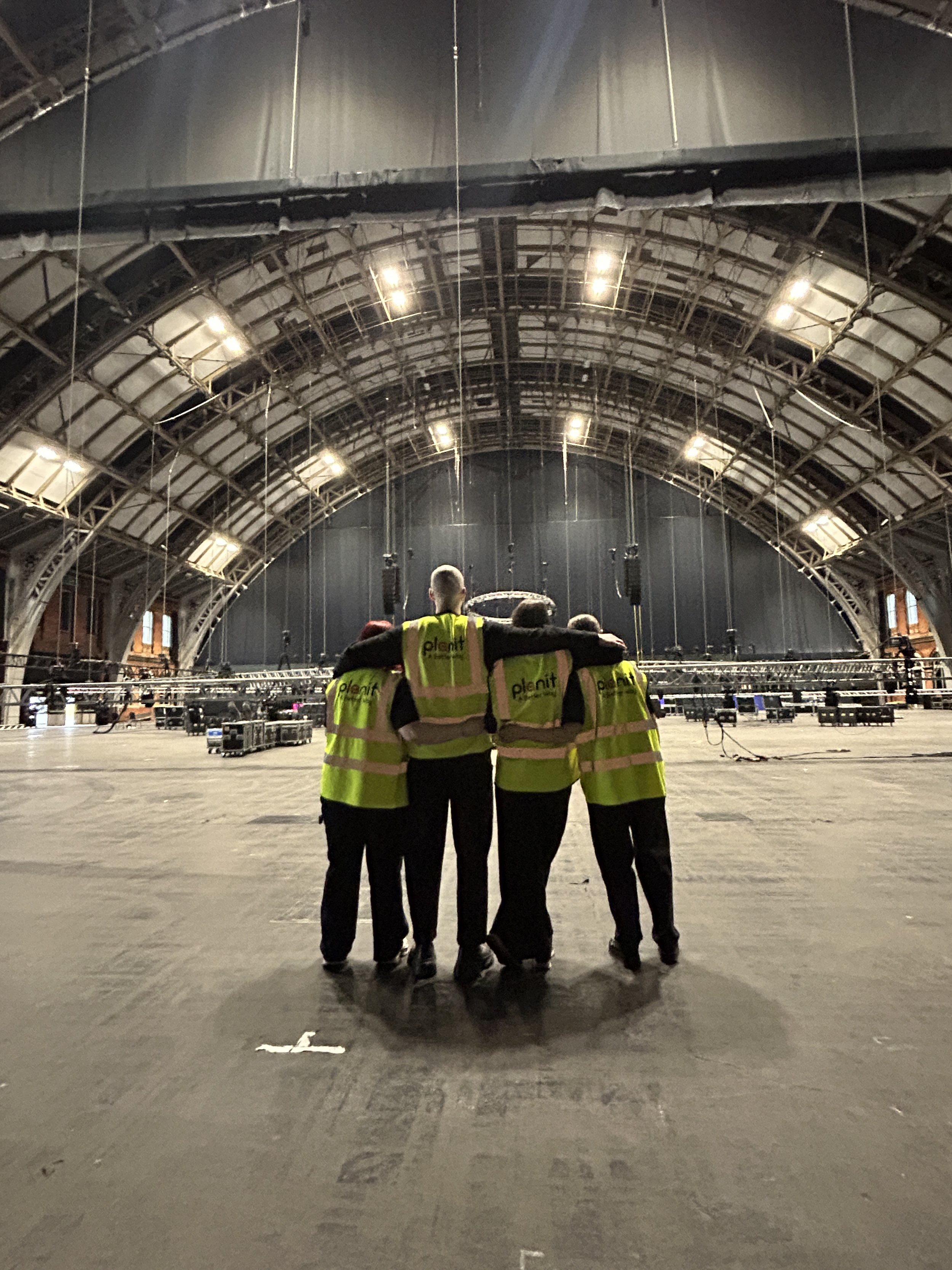 Five people wearing yellow safety vests with the word 'Planet' on the back are walking in an empty, large indoor space with a curved ceiling structure, overhead lights, and some scaffolding or equipment.