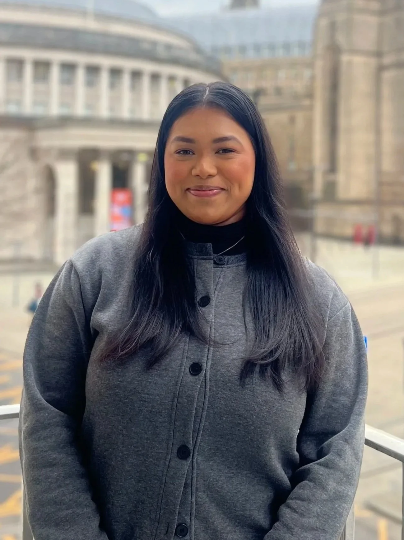 A woman with long black hair wearing a gray jacket standing outdoors with a large classical building in the background.