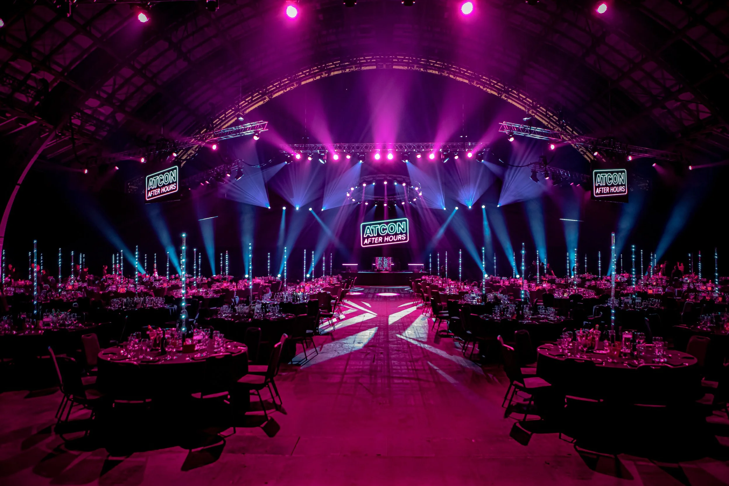 A large event venue with round tables set for dining, vibrant pink and purple lighting, a stage with multiple screens displaying the logo "ATCON After Hours," and an arched ceiling structure.