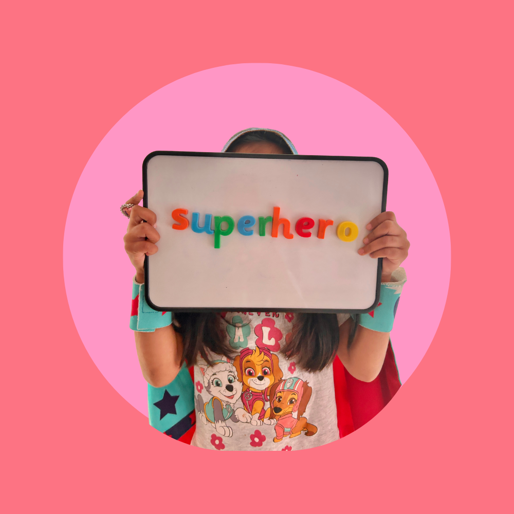 Child holding a sign that spells out 'Superhero' with colorful magnet letters, standing against a pink background with a darker pink circle behind her.