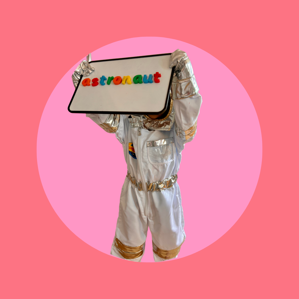 Child dressed in an astronaut suit holding a sign that says "astronaut" with colorful letters, against a pink background.