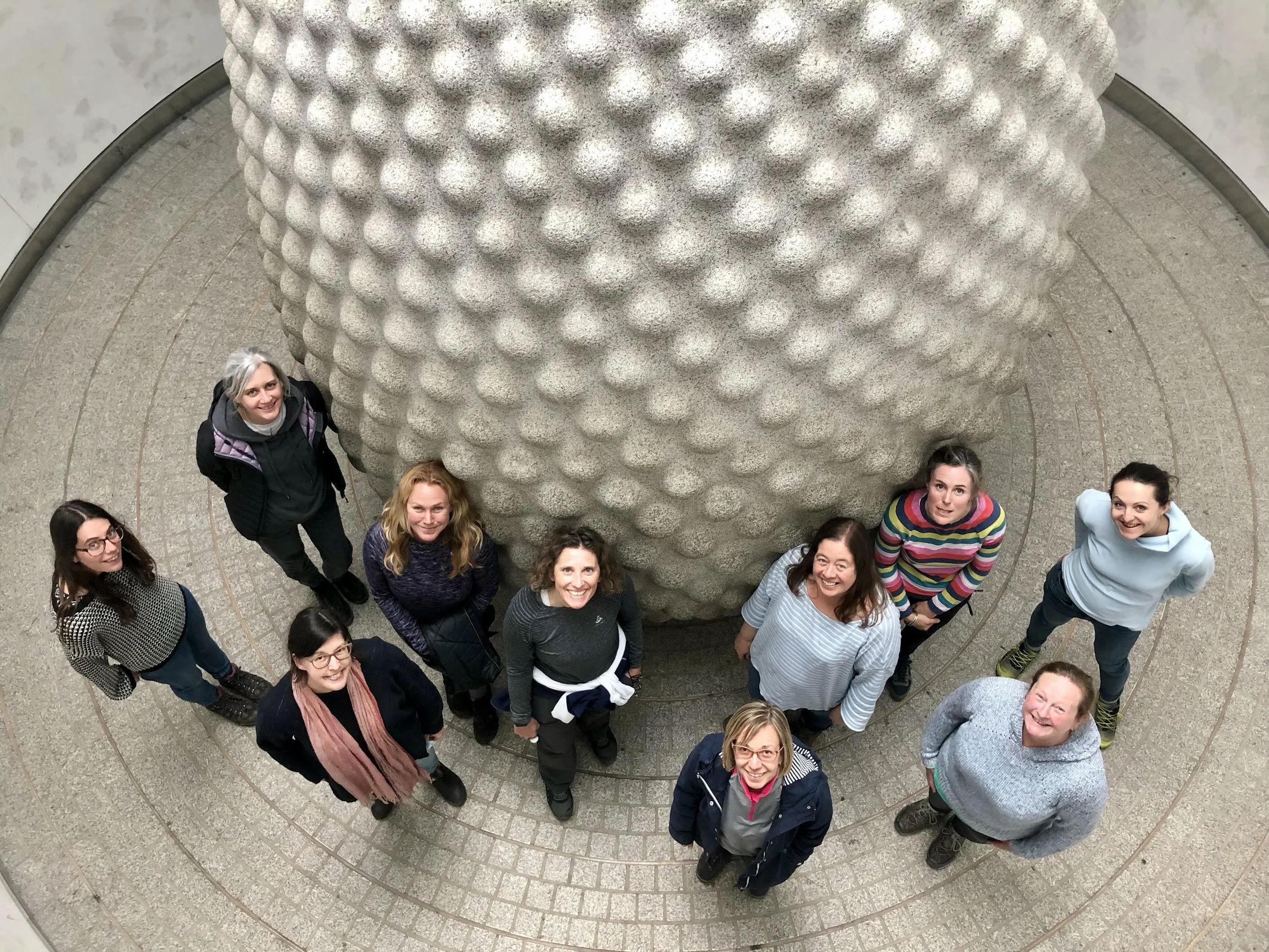 A group of ten women looking up at the camera inside a circular area near a large, textured, egg-shaped sculpture.