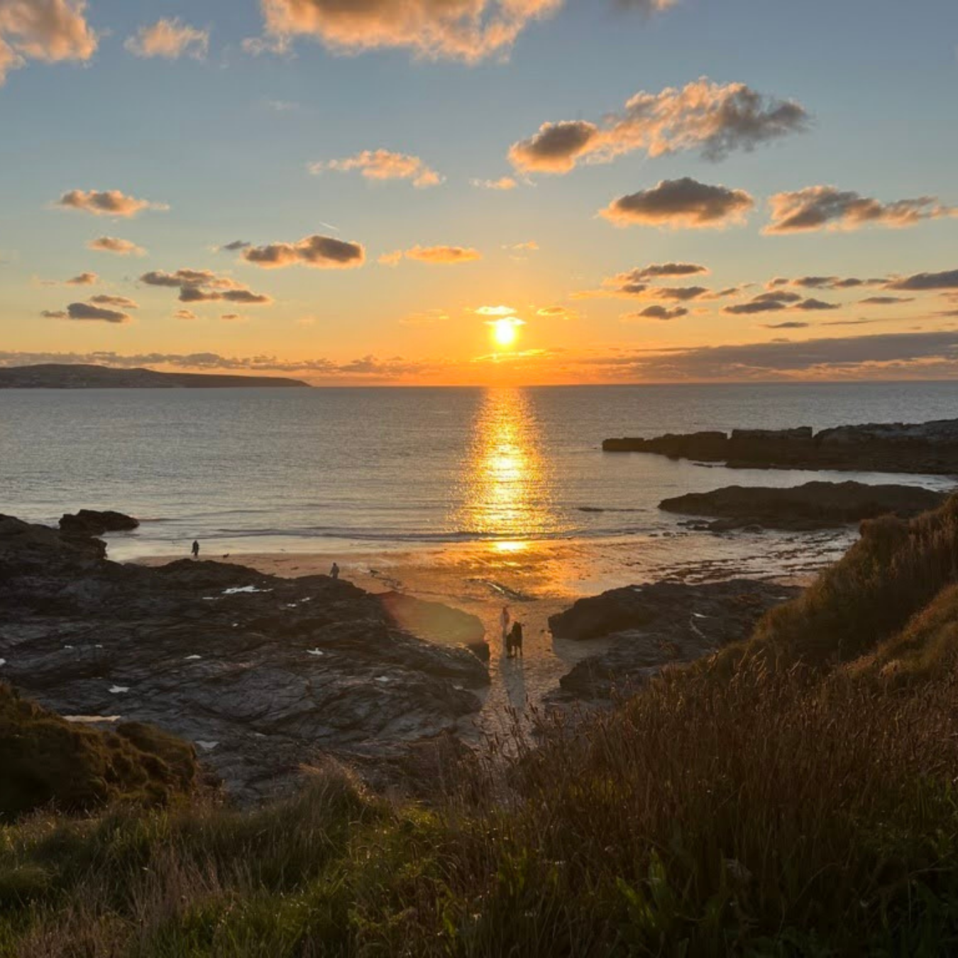 Sunset over a beach with rocks and greenery in the foreground, and a few people walking along the shoreline.