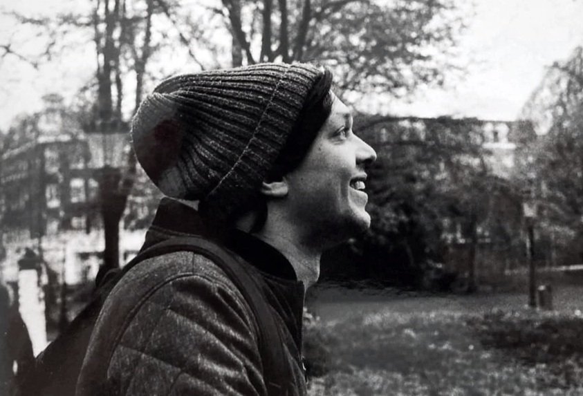 Black and white photo of a smiling man wearing a beanie and a jacket, standing outdoors with trees and buildings in the background. He seems creative, a composer, friendly, good to work with.
