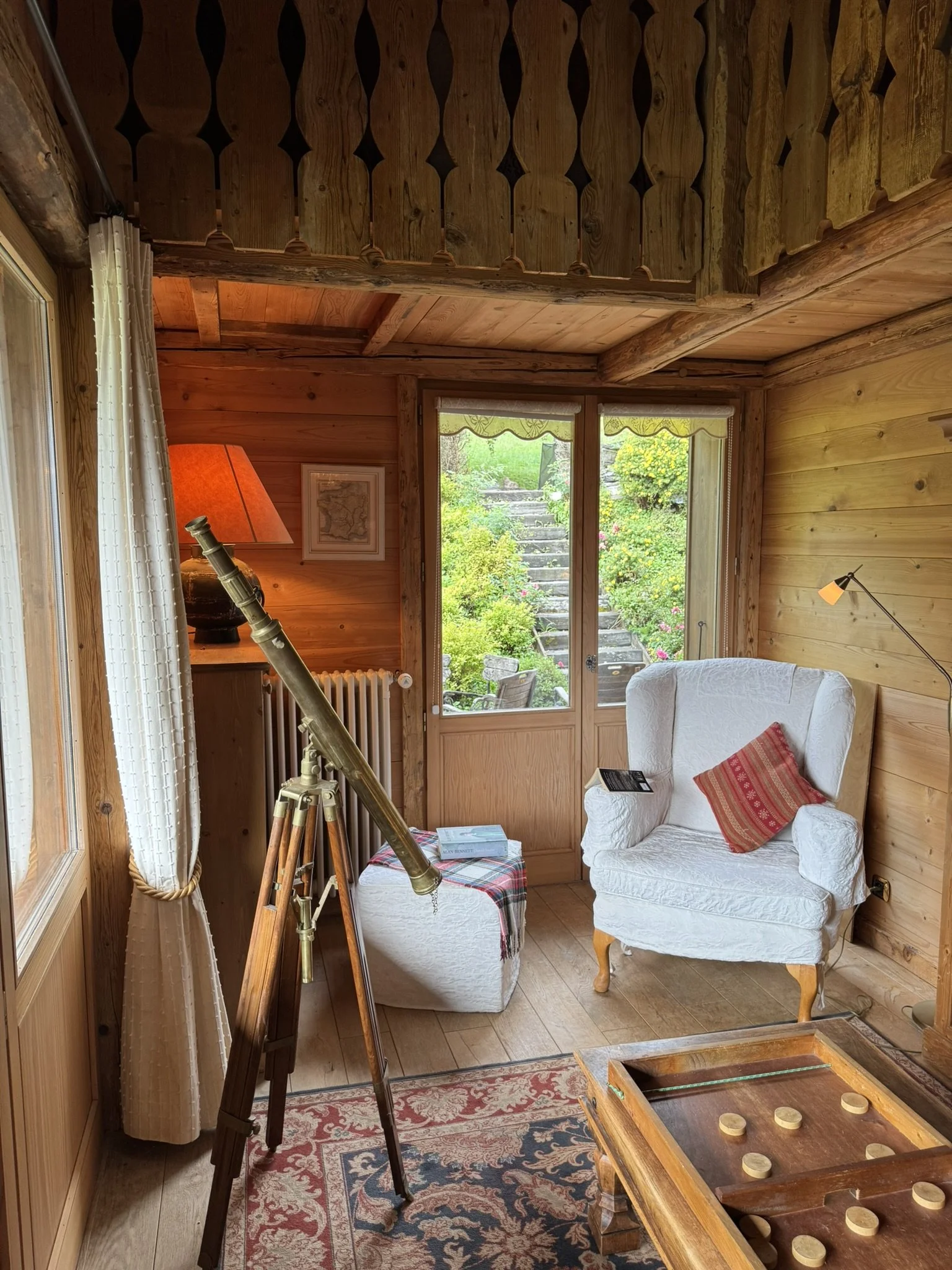 A cozy wooden interior room with a telescope near a window, white armchair with a red pillow, a side table with books, a lamp, and a view of garden stairs outside.
