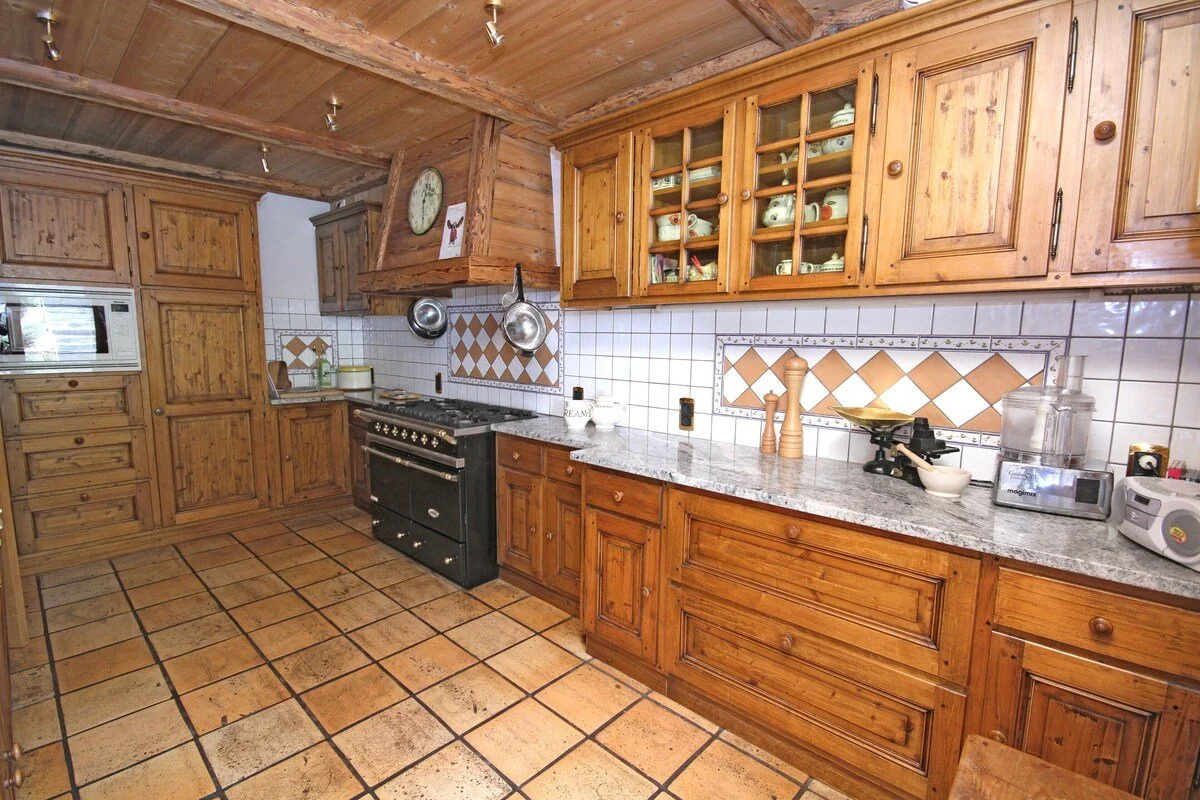 A rustic kitchen with wooden cabinets, a tiled floor, and a granite countertop. There is a black stove, a microwave, and various kitchen utensils and appliances on the counter. Hanging pans are above the stove, and dishes are stored in glass-front cabinets.