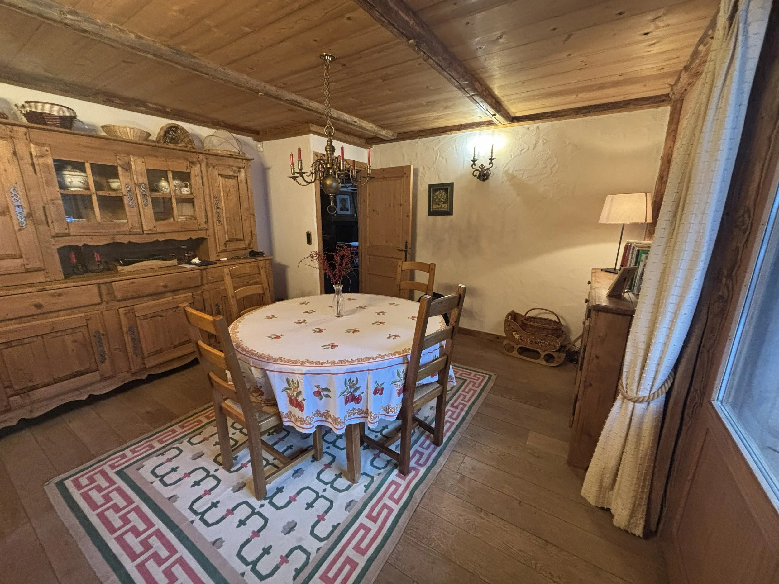A rustic dining room with a round wooden table surrounded by five matching wooden chairs. On the table are a vase with red berries, a tablet, a cup, some papers, and a small device. In the background, there is a large wooden hutch with glass doors and baskets on top, a chandelier overhead, and a partially open wooden door leading to a living room with a fireplace and a red sofa.