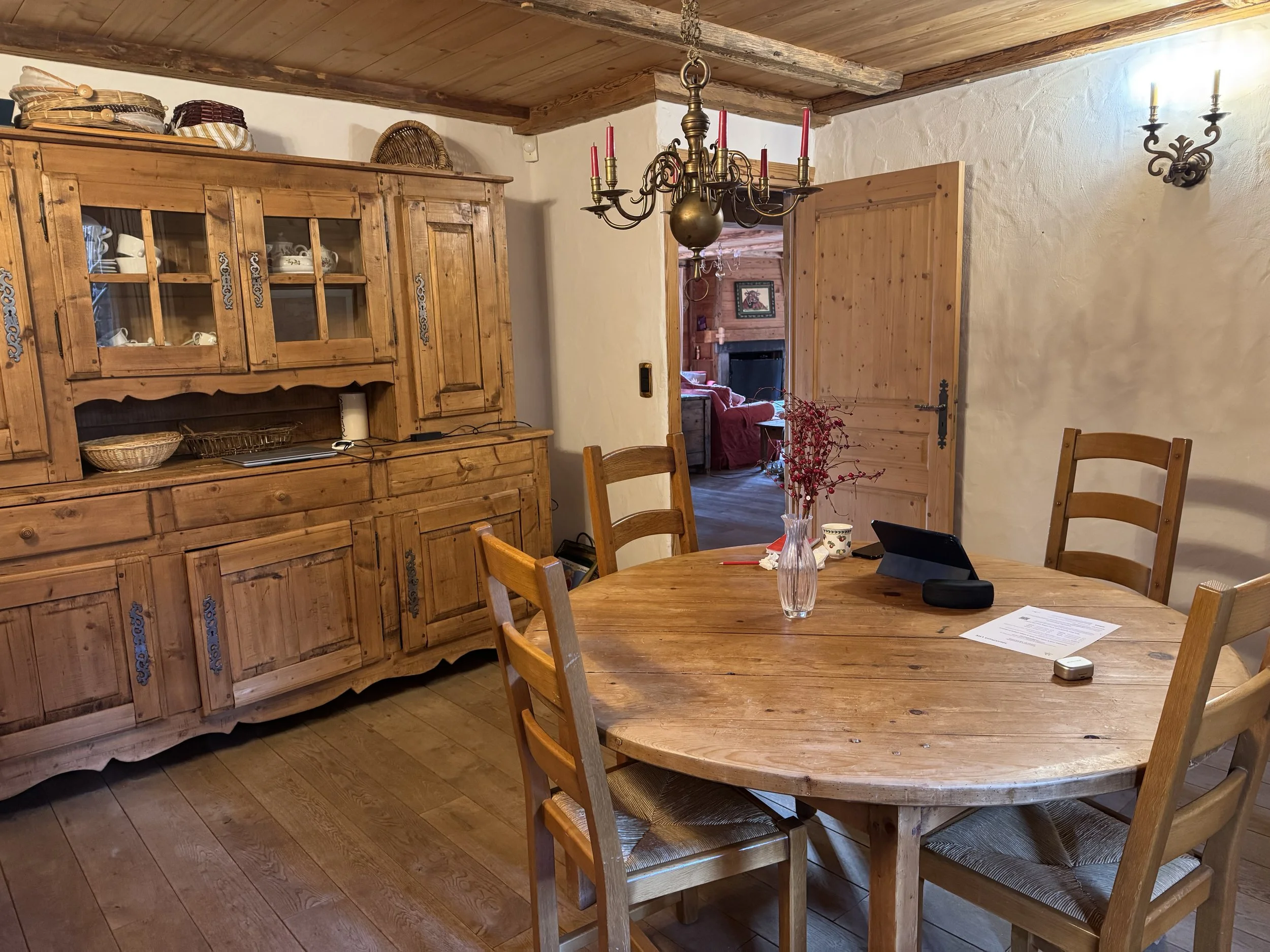 A rustic dining room with a round wooden table surrounded by five matching wooden chairs. On the table are a vase with red berries, a tablet, a cup, some papers, and a small device. In the background, there is a large wooden hutch with glass doors and baskets on top, a chandelier overhead, and a partially open wooden door leading to a living room with a fireplace and a red sofa.